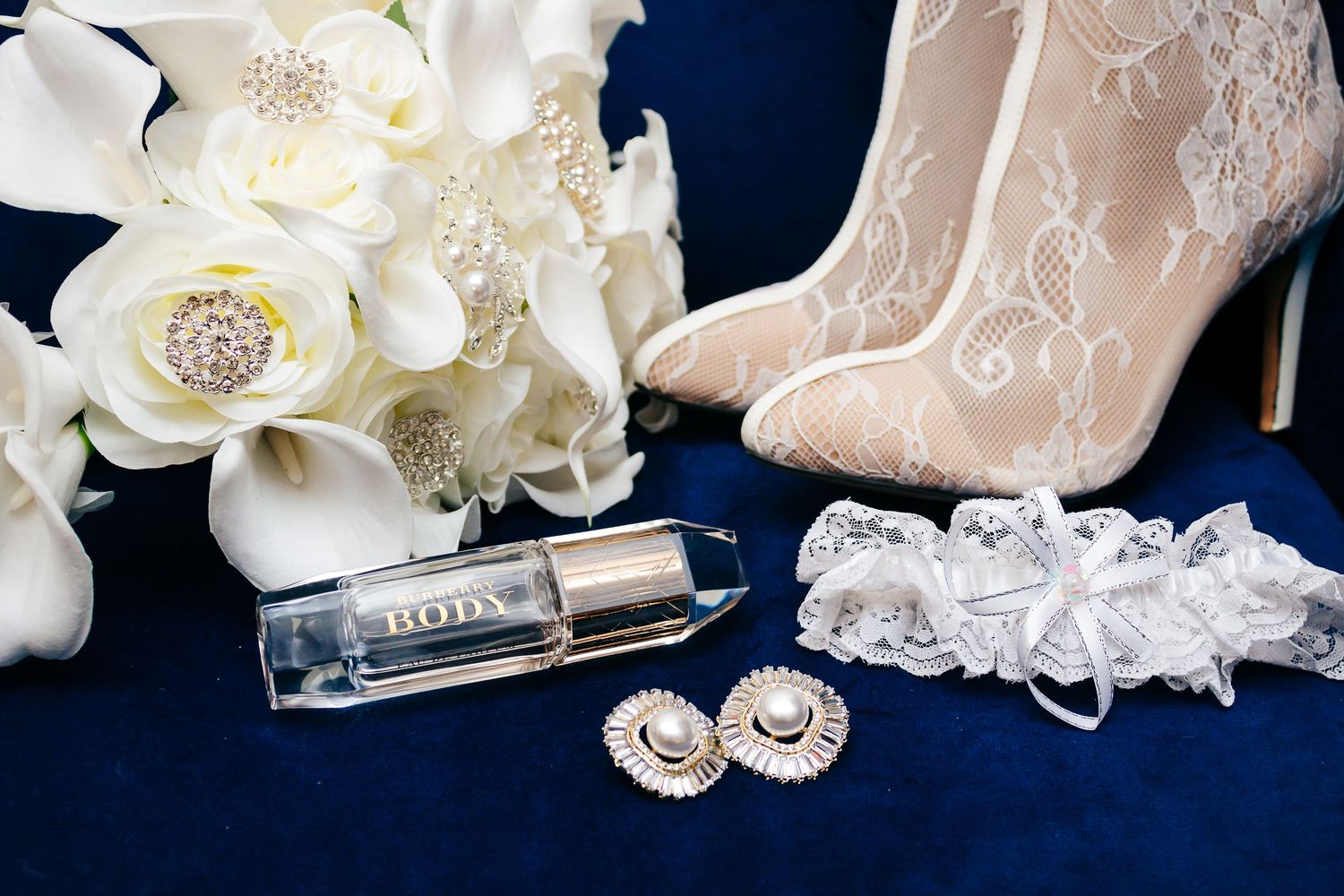 Wedding accessories including lace shoes, white bouquet, perfume and jewelry arranged on dark blue velvet background.