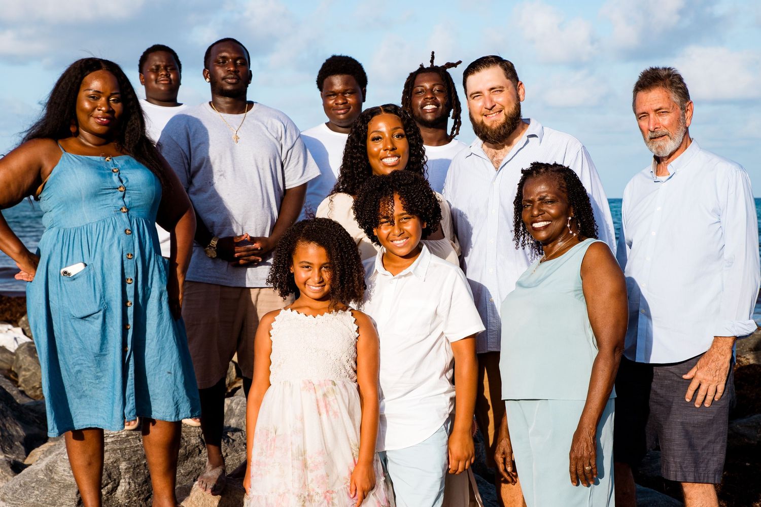 A diverse group of people wearing casual clothing pose together outdoors against a bright blue sky background.