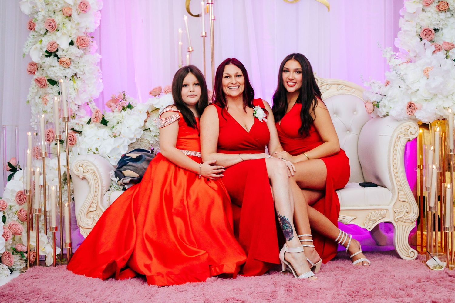 Wedding party in red dresses poses by floral wall installation with pink lighting.