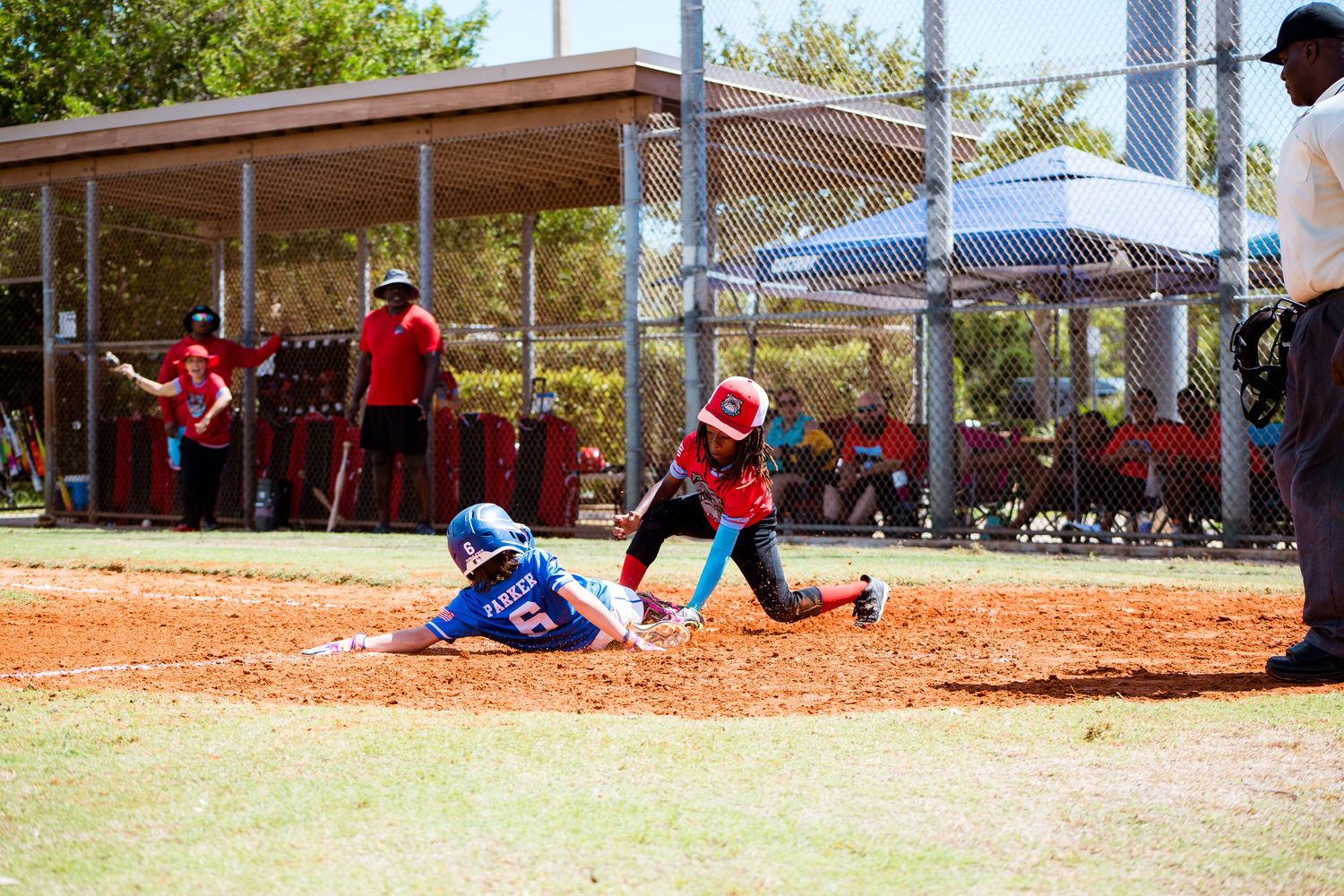 Youth baseball game underway on a sunny day with covered dugouts visible in the background.