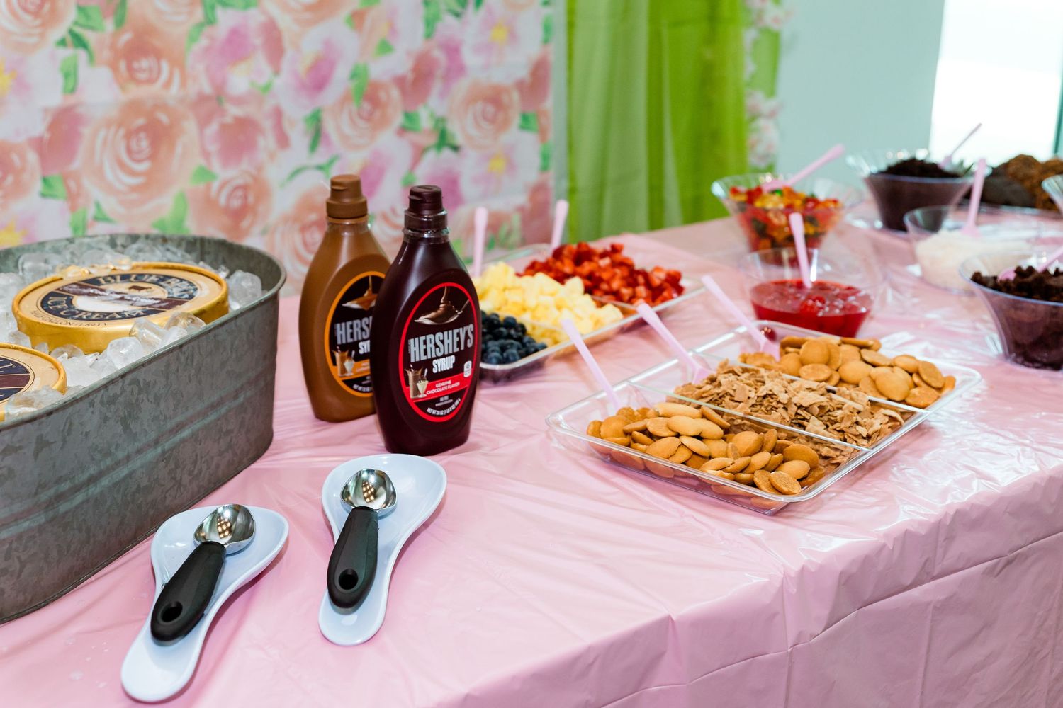 Close-up of dessert table with chocolate sauce bottles, scoops, and snack platters on pink tablecloth.