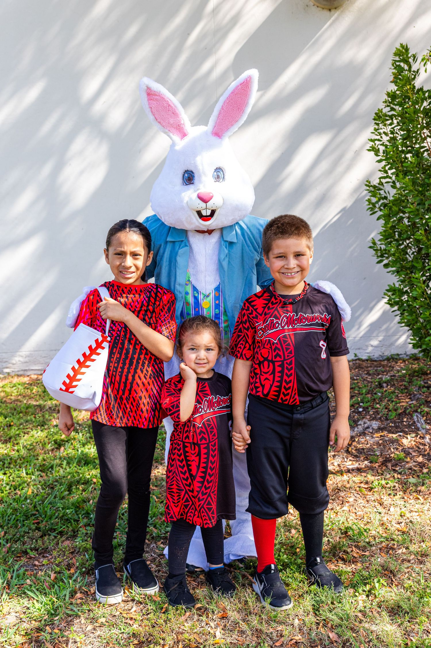 Three children pose with Easter Bunny mascot outdoors, wearing matching red tribal print baseball uniforms.