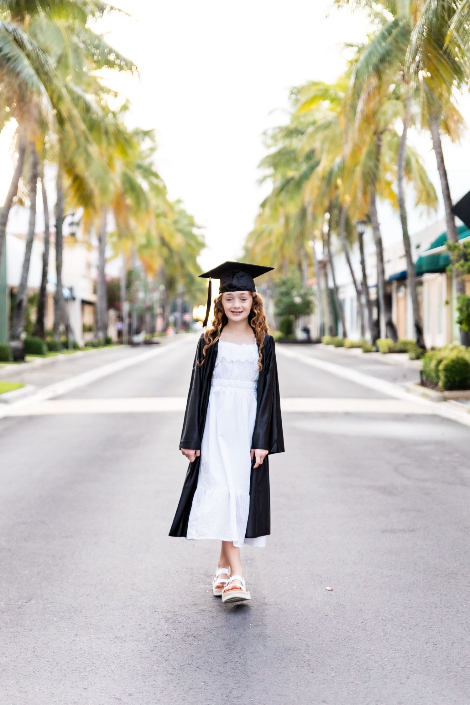 Young girl in black graduation cap and gown over white dress walks down palm-lined street, smiling.
