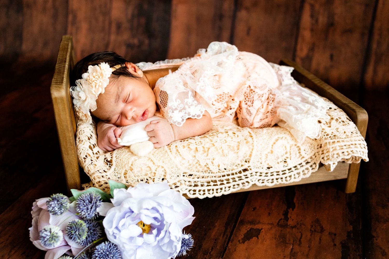 Sleeping newborn baby in lace outfit on tiny wooden bed with floral headband and blue flowers on rustic wood background.