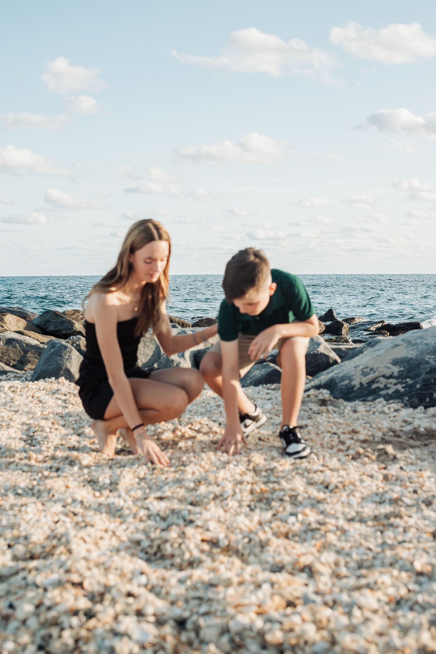 Two people crouched on a rocky beach searching for seashells at sunset along the ocean shore.