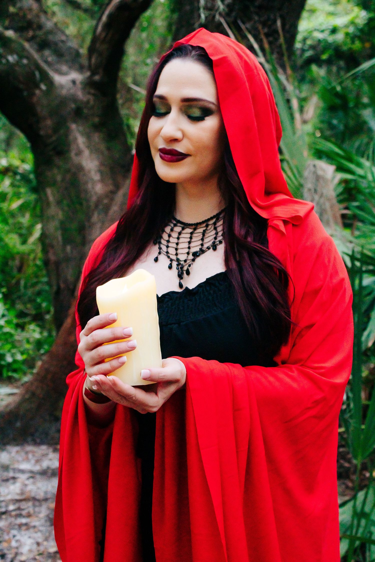 A mysterious figure in a red cloak holds a candle while standing in a misty forest setting.