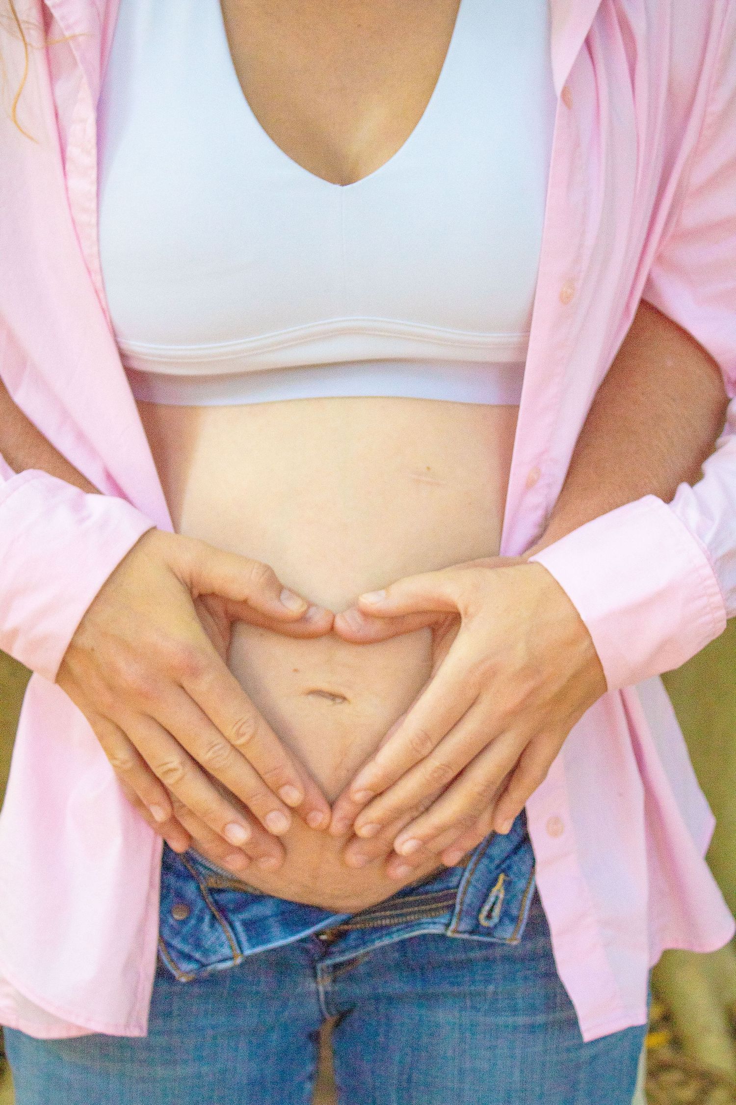 Woman in pink cardigan and jeans making heart shape with hands over pregnant belly.