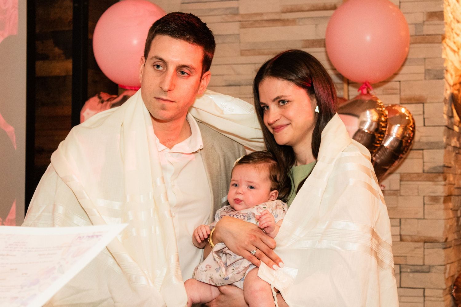 A couple holding an infant at a celebration with pink and rose gold balloon decorations.