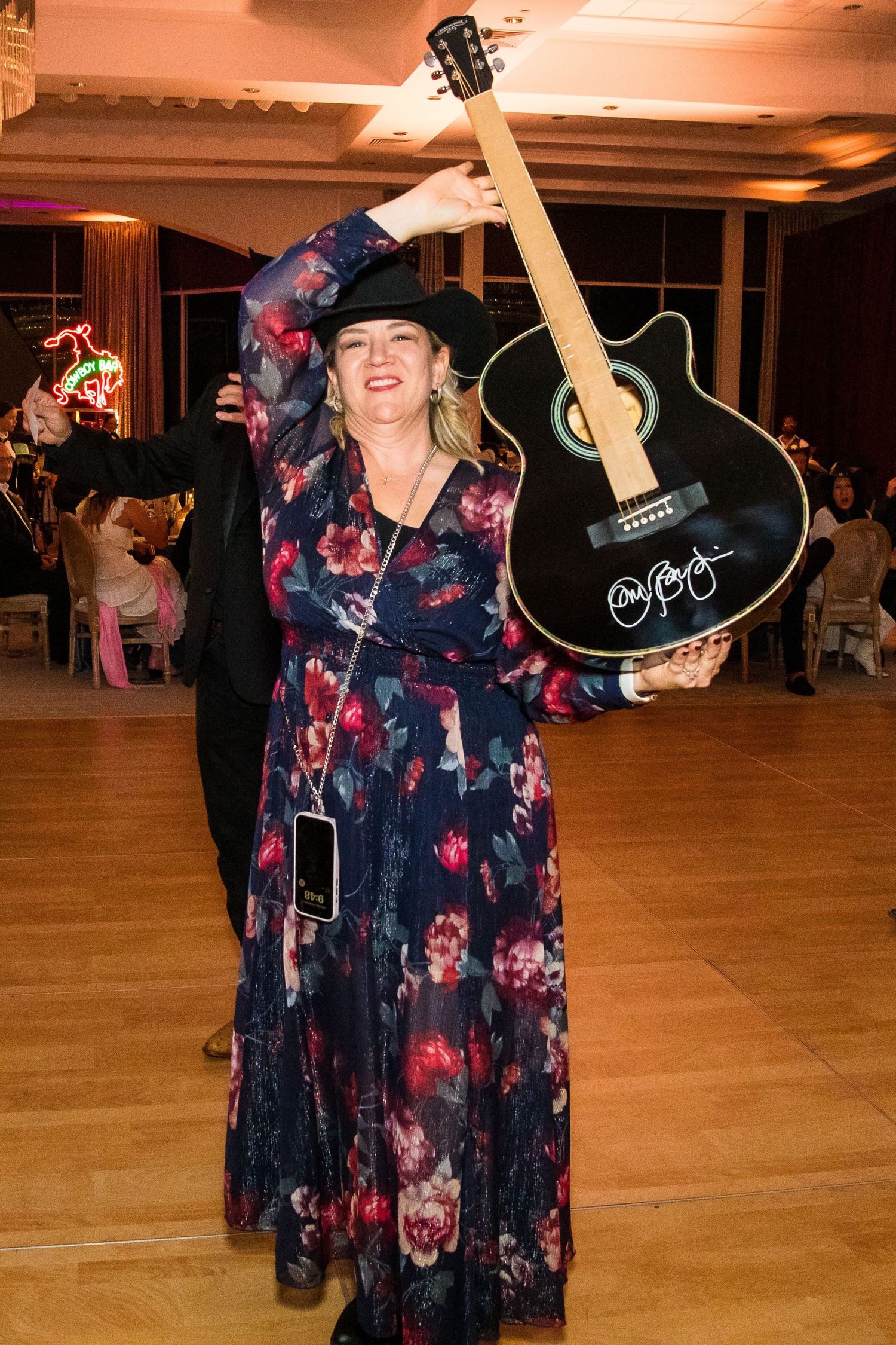Person in floral dress holding up black acoustic guitar at formal event.