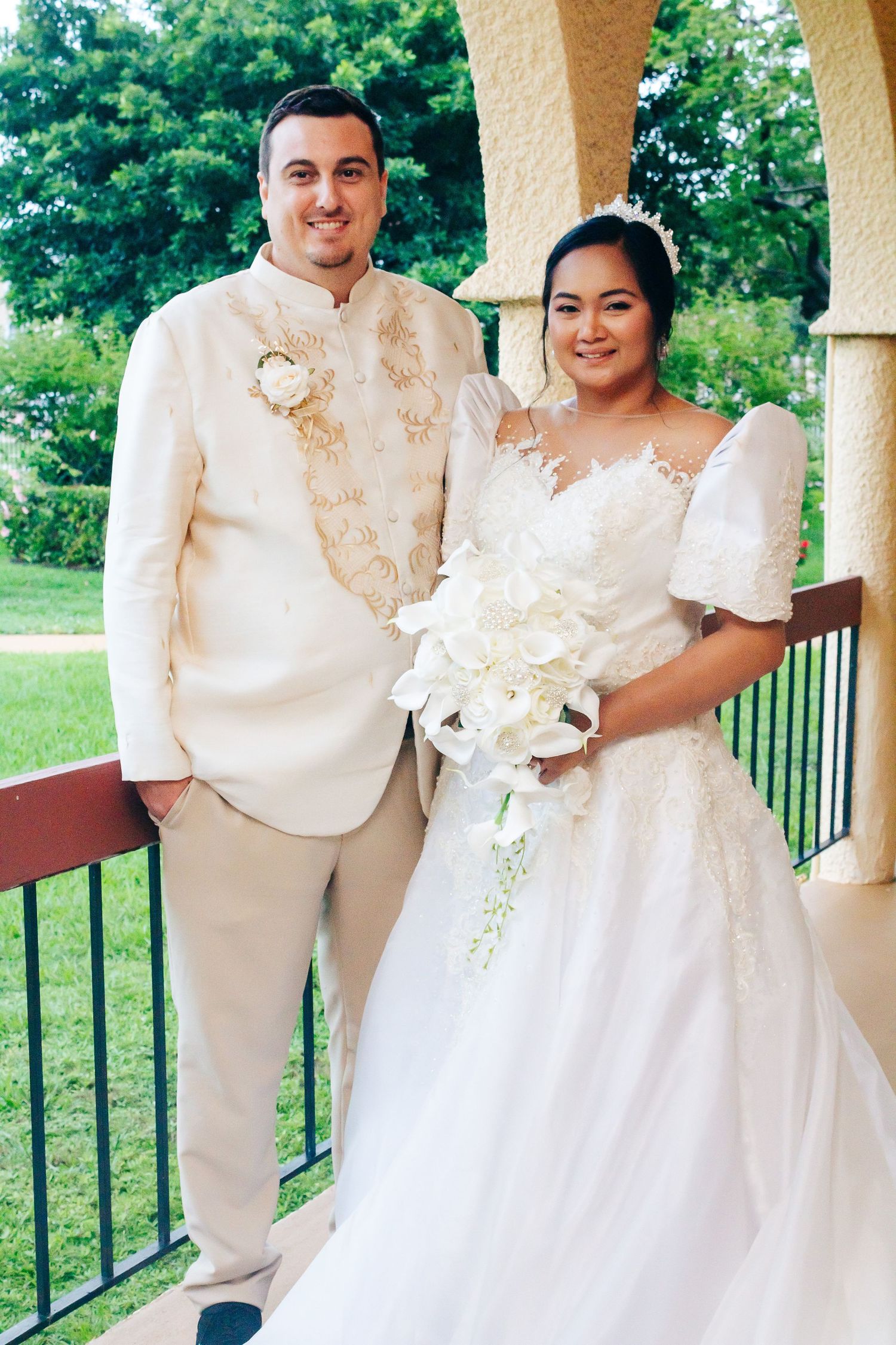 Newlywed couple poses outdoors on a porch in white traditional wedding attire with a floral bouquet.