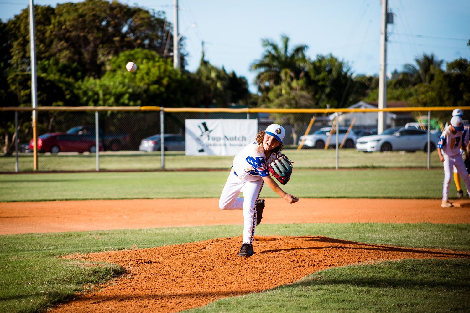 Baseball pitcher in white uniform throwing a pitch from the mound during a game on a sunny field with palm trees.