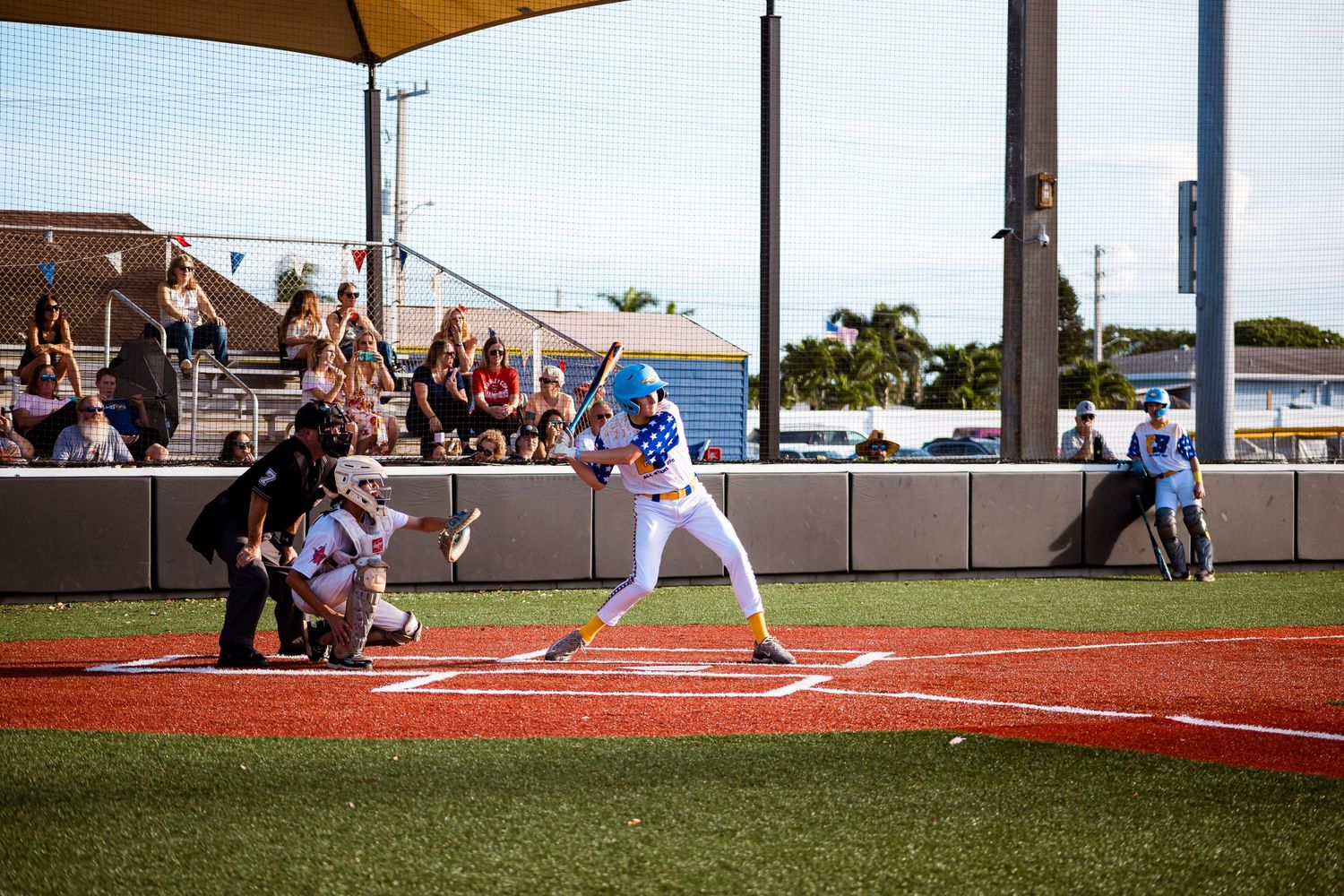 Baseball player in blue uniform at bat on sunny day while fans watch from covered bleachers at outdoor stadium.