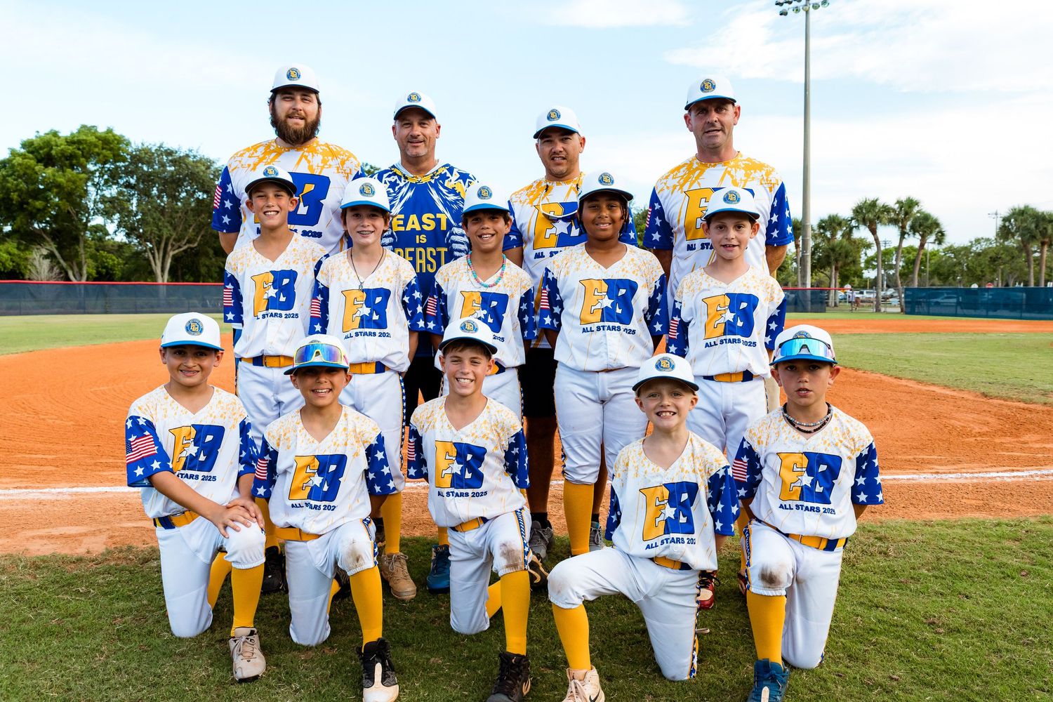 Youth baseball team poses together in blue and white uniforms on baseball field with coaches standing behind them.