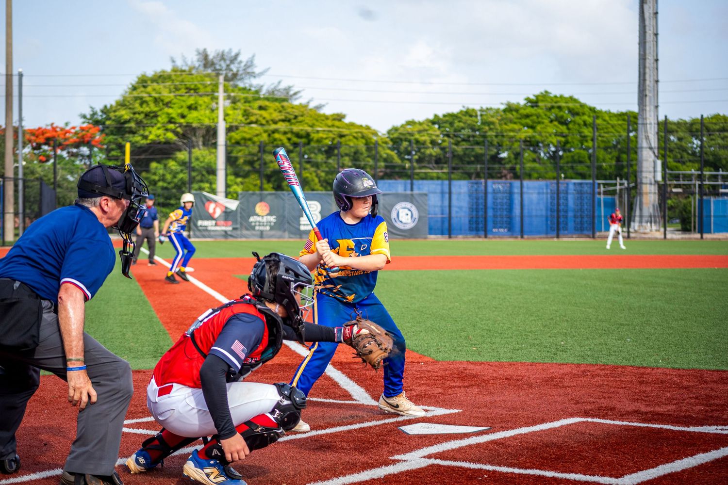 Baseball batter in blue and yellow uniform prepares to swing as catcher and umpire crouch behind home plate.