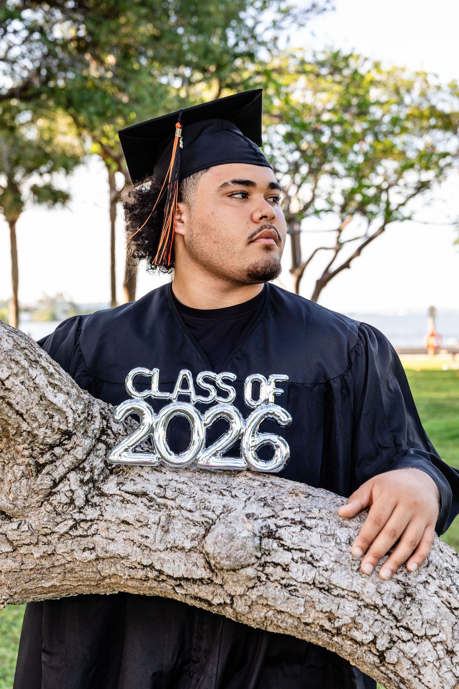 Young man in black graduation cap and gown holding Class of 2026 balloon sign while leaning on tree outdoors.
