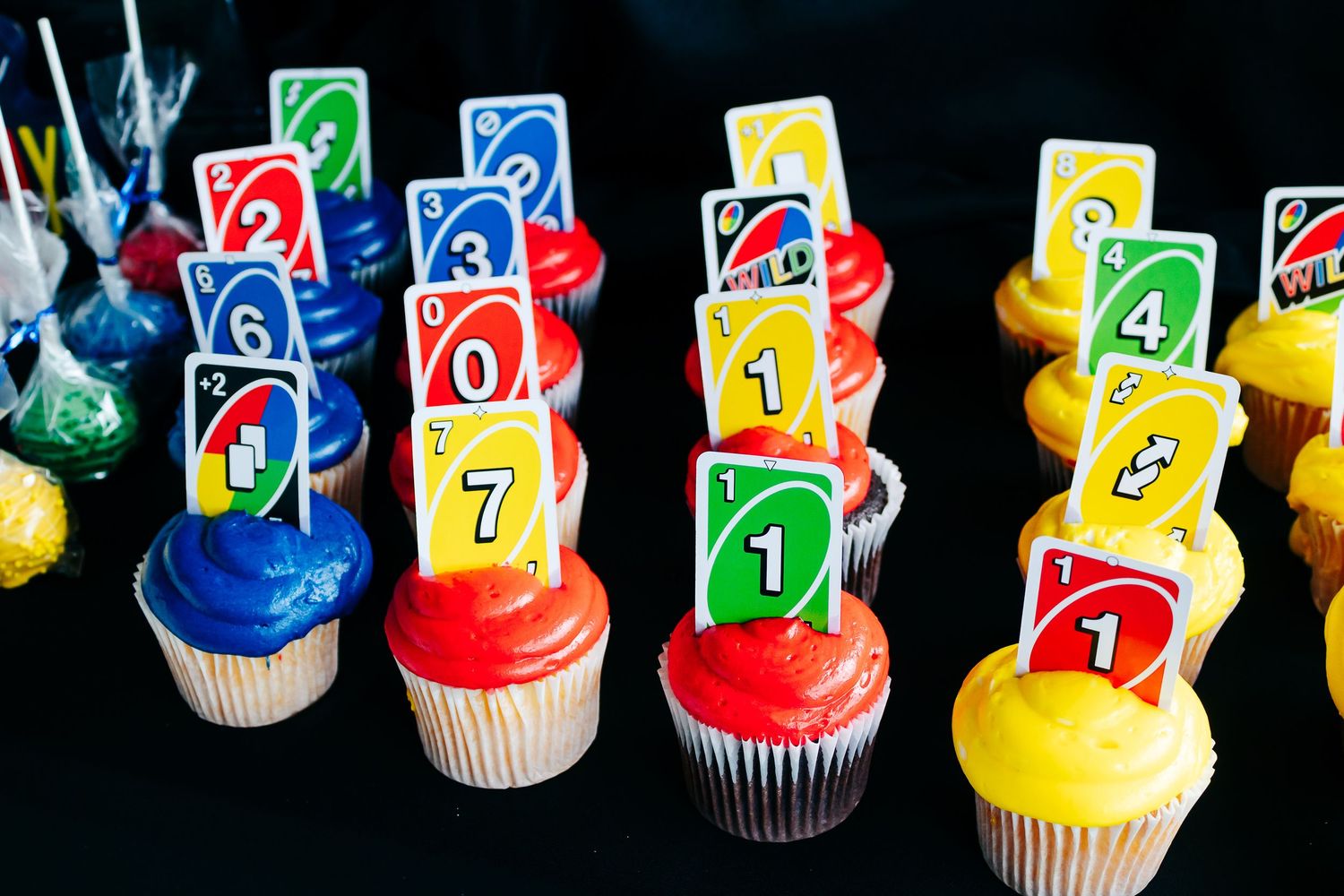 Colorful cupcakes decorated with UNO card themed toppers arranged on black background.