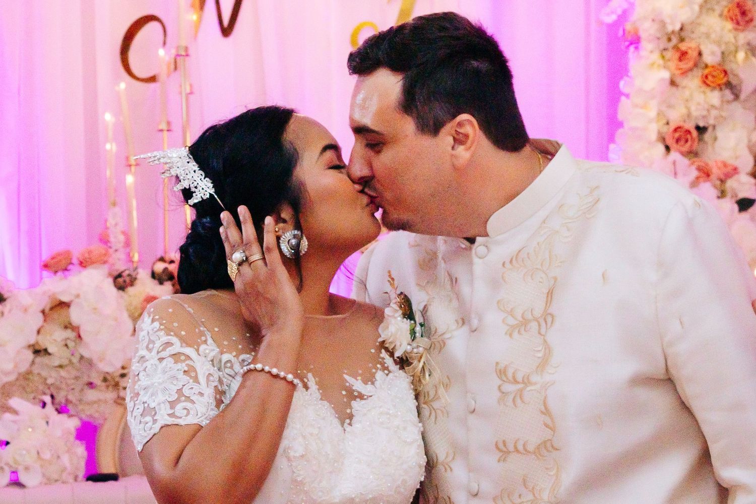 A bride and groom share intimate kisses at their wedding reception with pink lighting and floral decor.