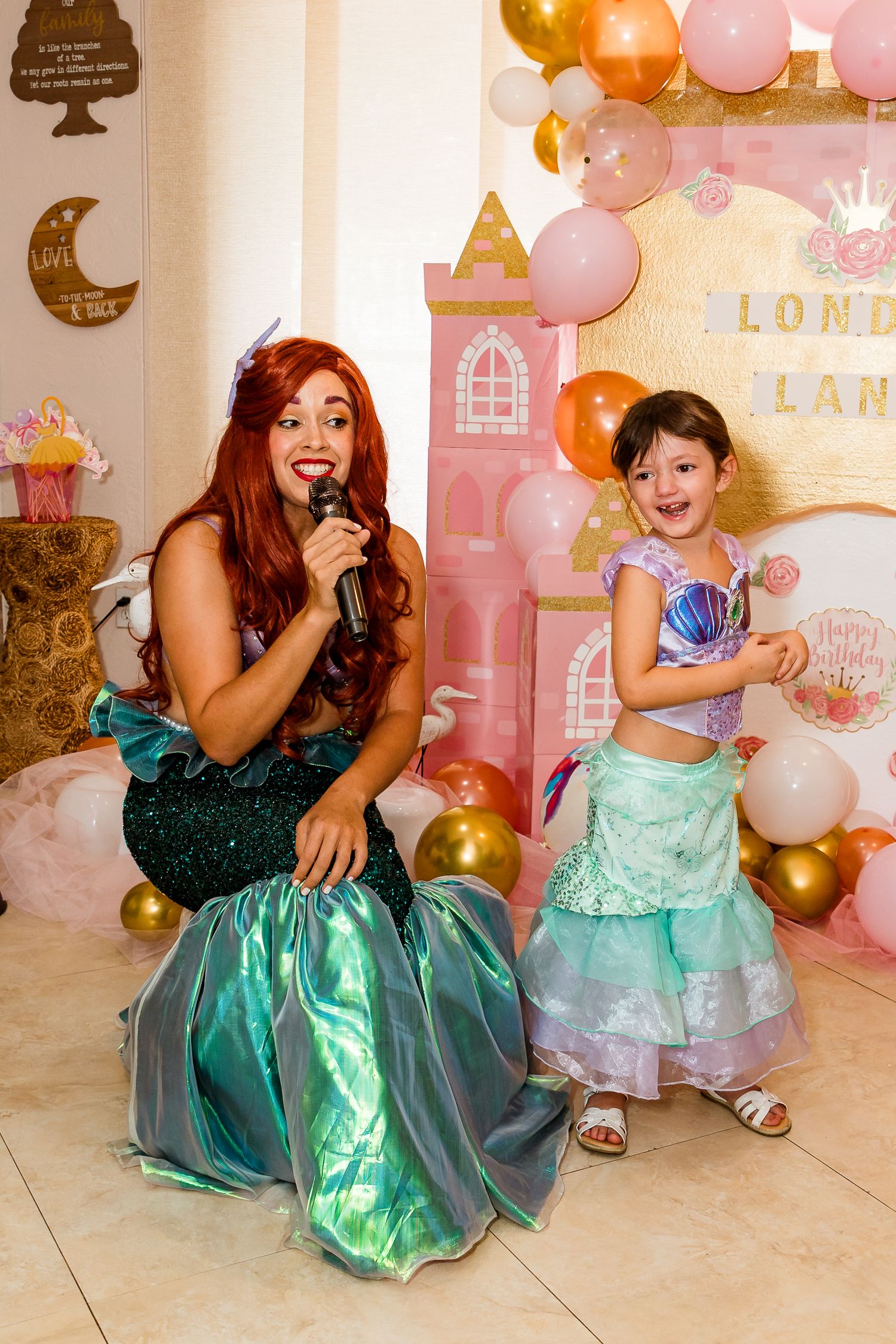 Princess-themed birthday party with matching green dresses and pink and gold balloon decorations at a castle backdrop.