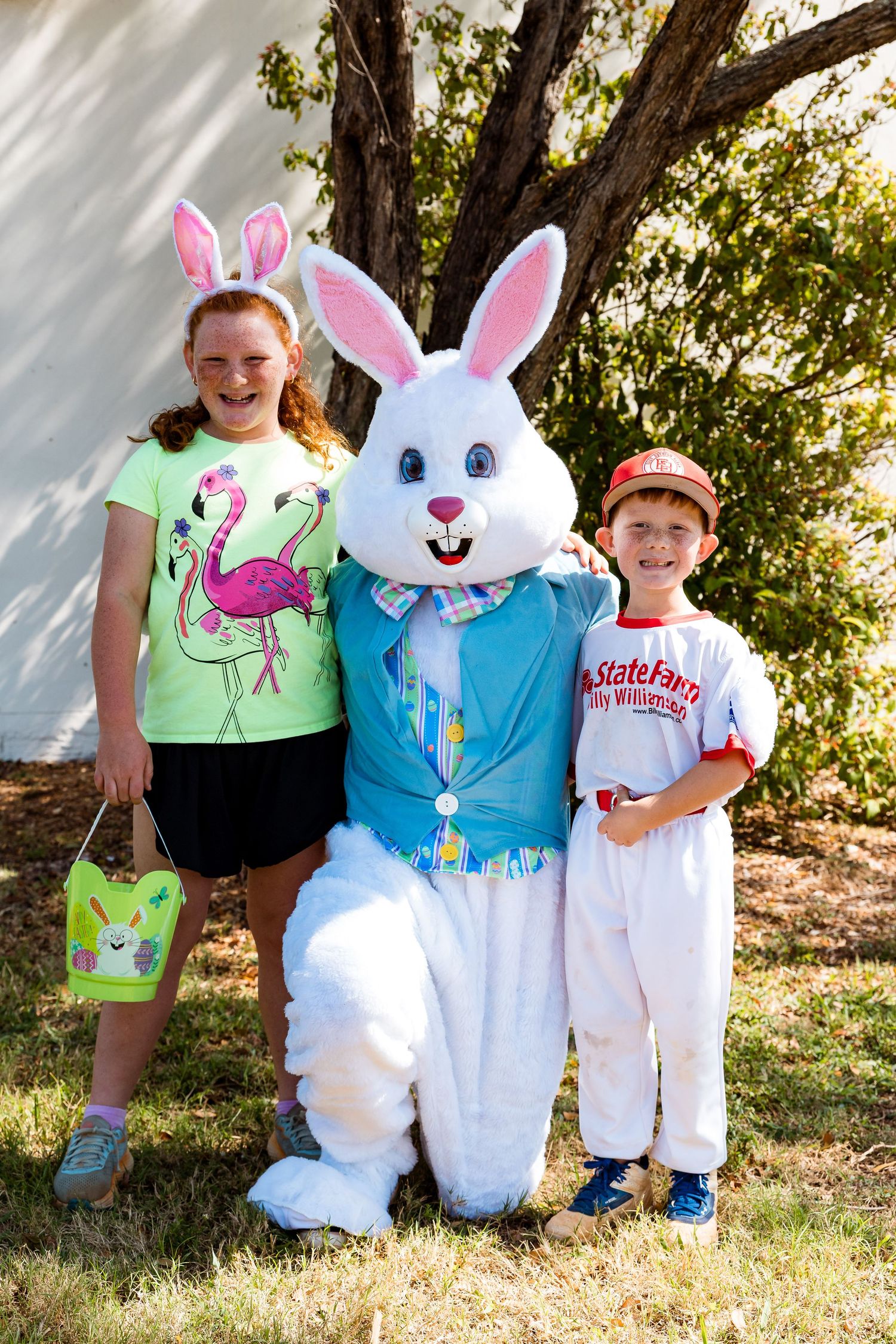 Two children pose outdoors with the Easter Bunny wearing a blue vest at a springtime celebration.