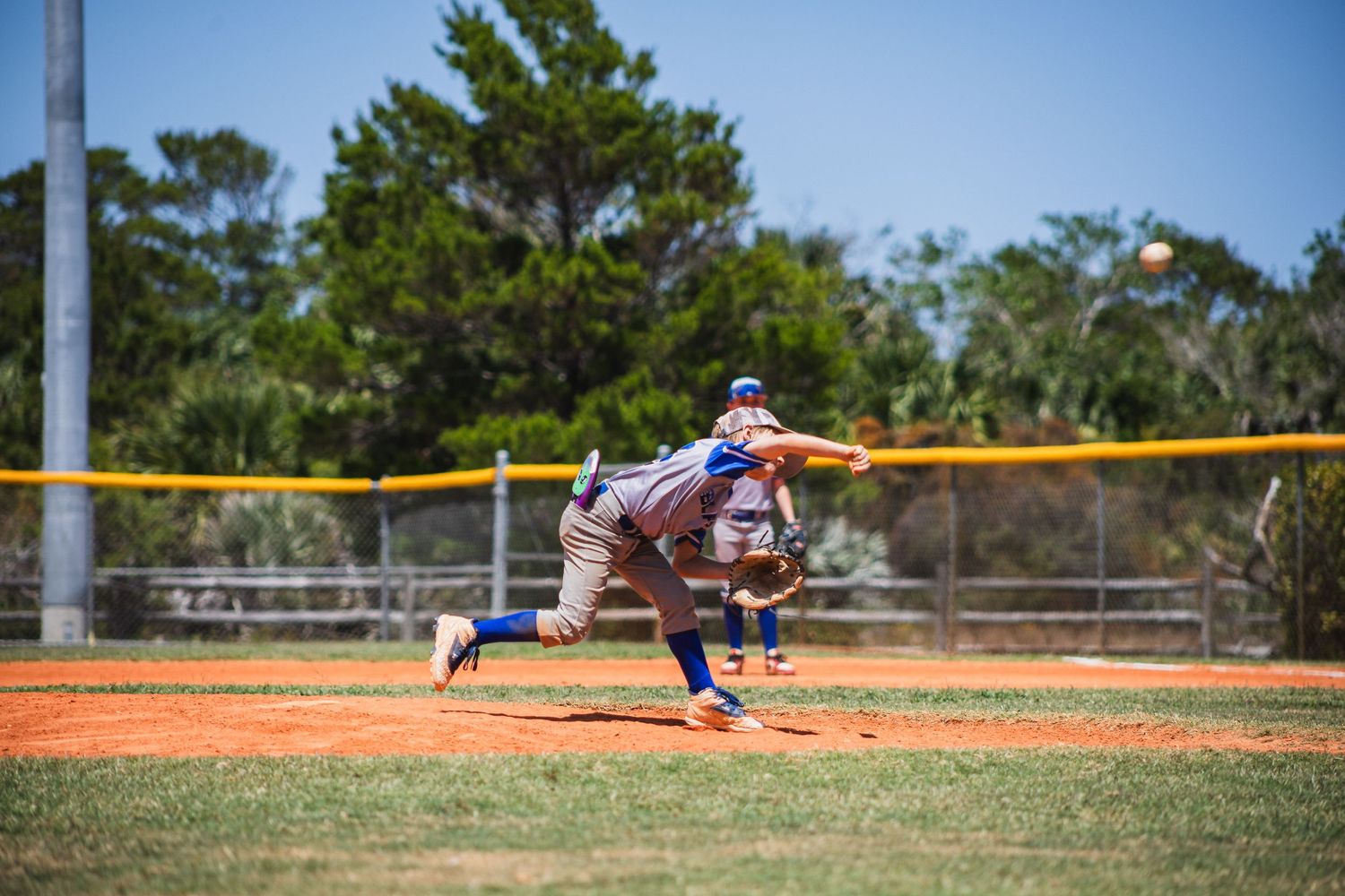 Baseball pitcher in mid-throwing motion on a sunny day at a baseball field with trees in the background.