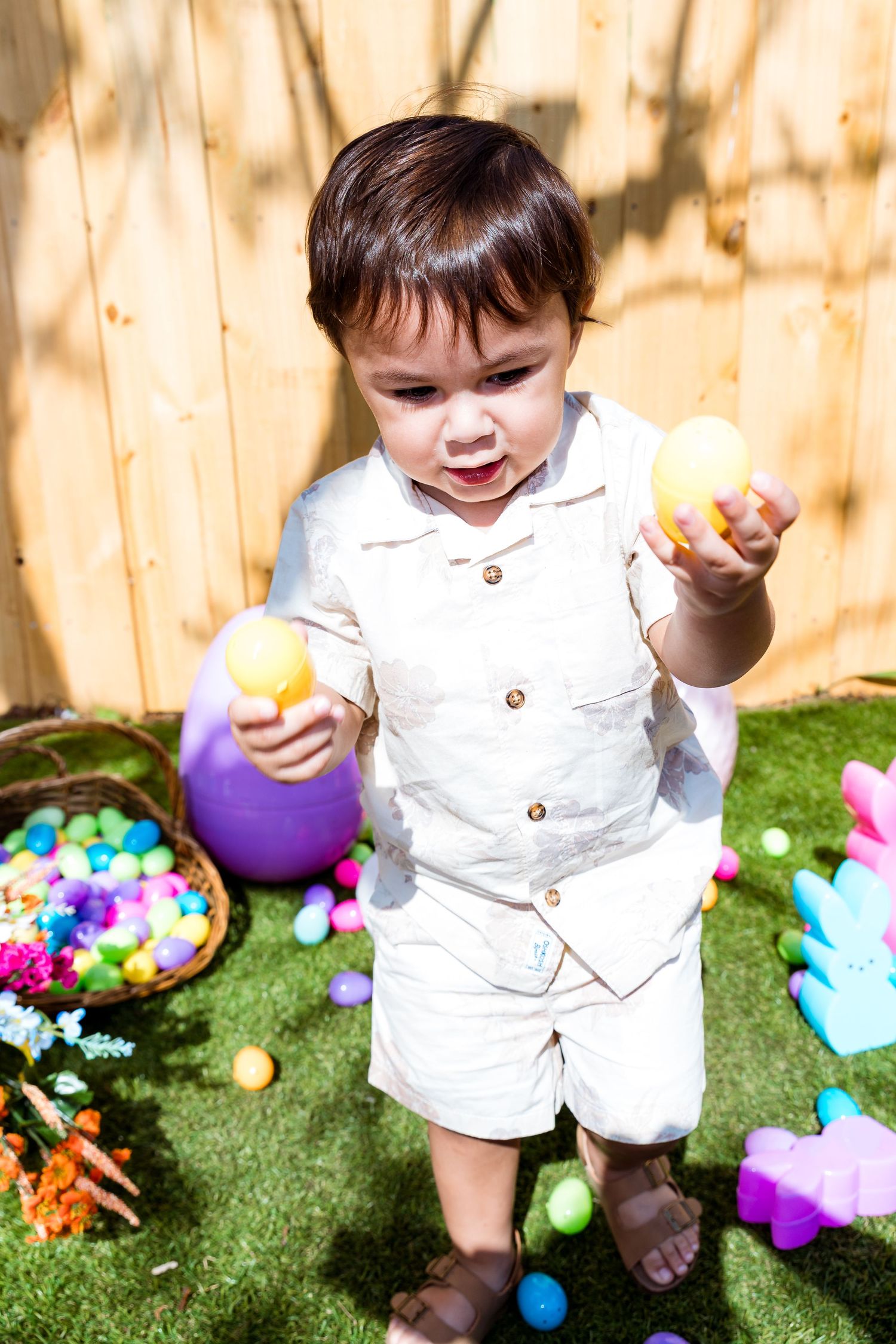 Child in white outfit holding Easter eggs with colorful scattered eggs and pastel decorations on grass.
