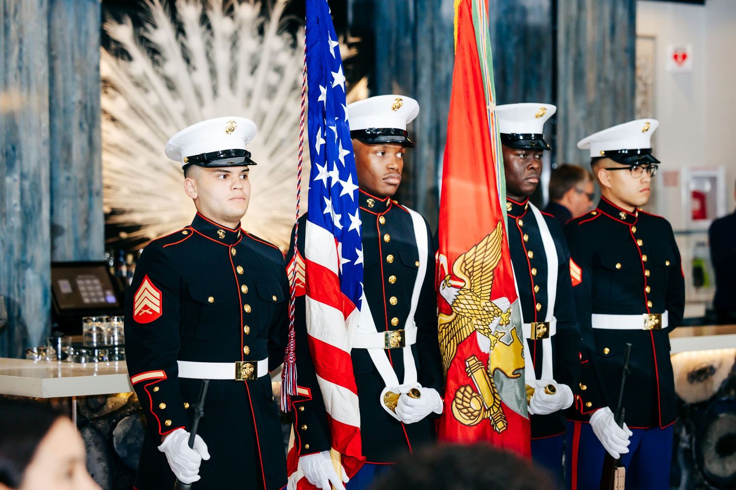 Military honor guard members in dress uniforms stand at attention holding American flags and service banners.
