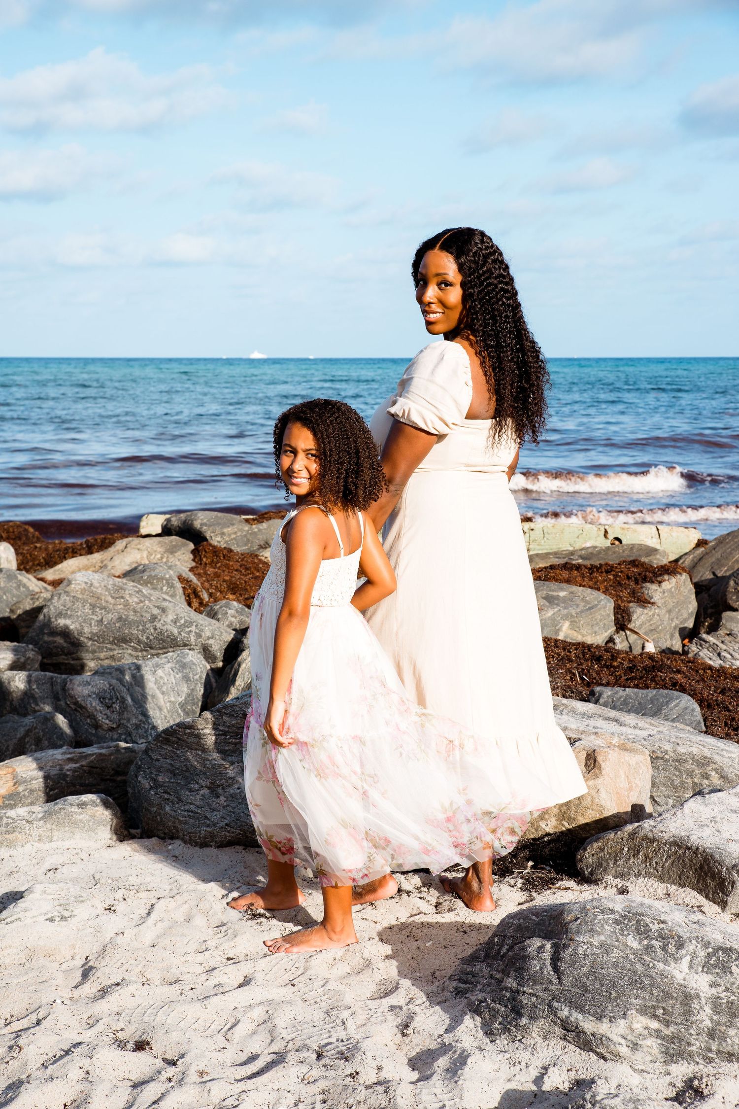 Two people in white dresses stand together on rocky beach landscape against blue ocean and sky background.