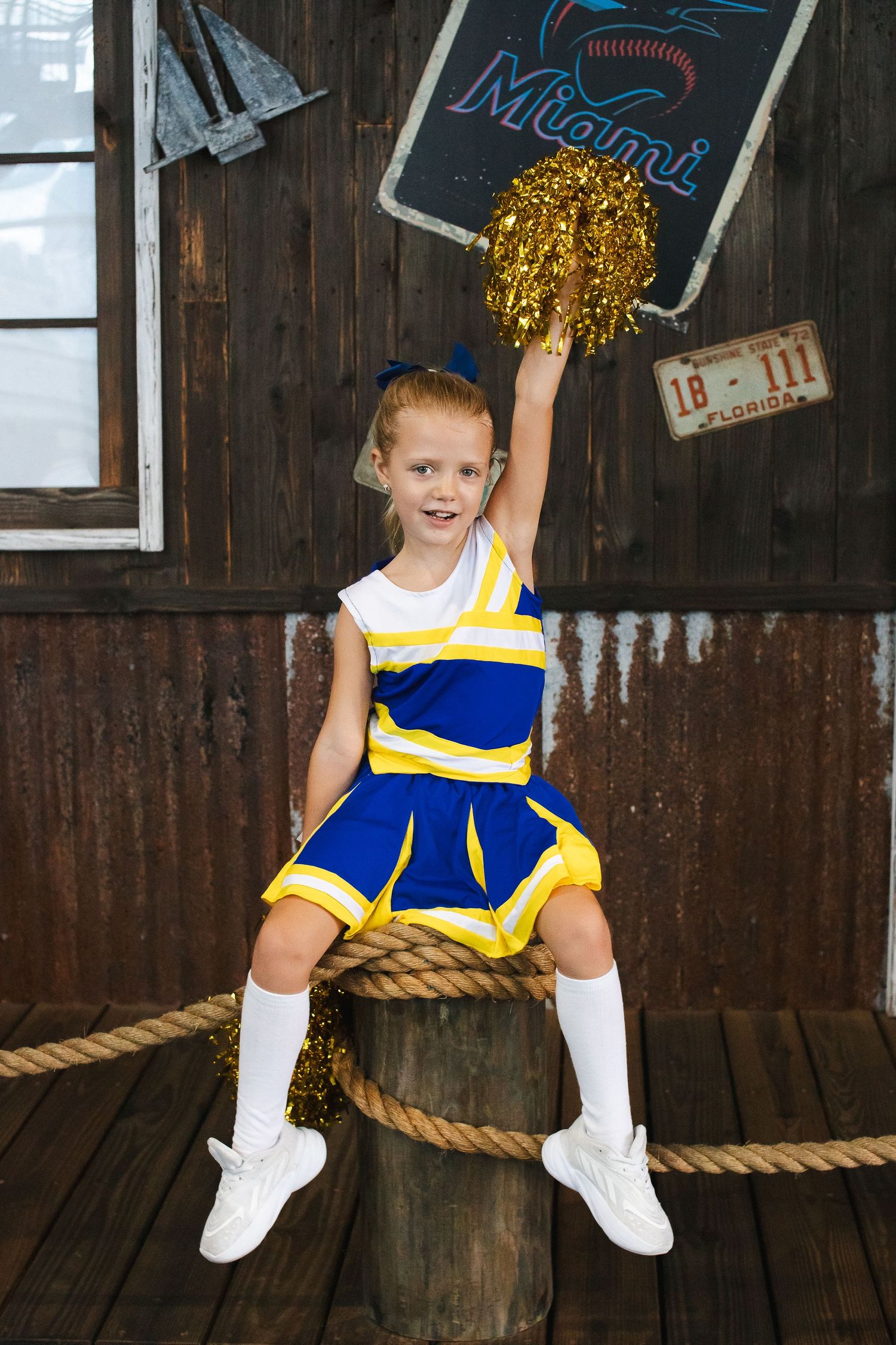 Young cheerleader in blue and yellow uniform holding gold pom poms while sitting on wooden prop.