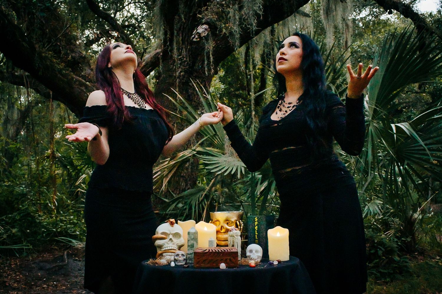 Two mystical figures in dark attire gesture dramatically beside a candlelit altar in a lush forest.