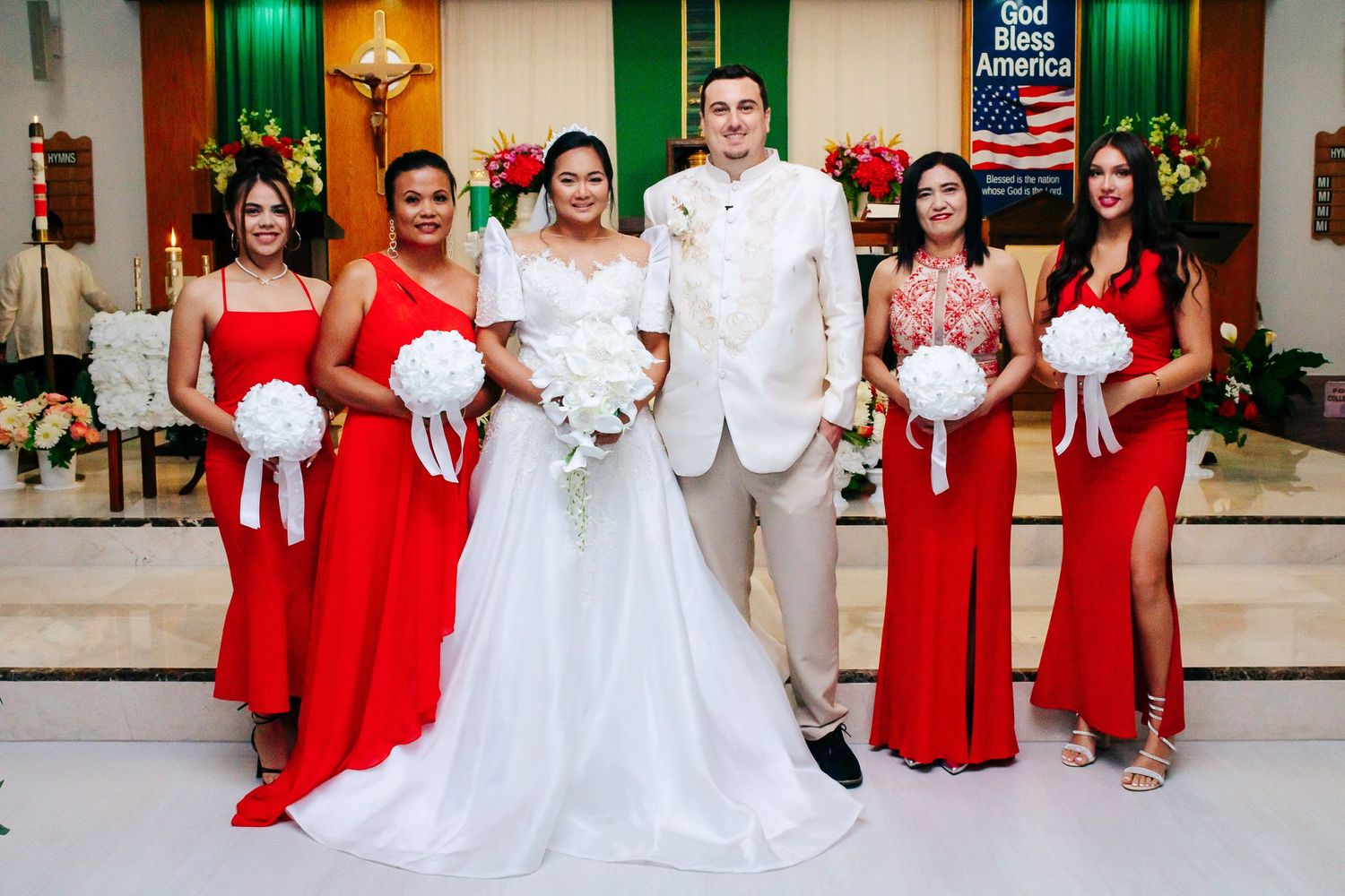 Bridal party in red dresses and groomsmen in red suits pose at church altar.