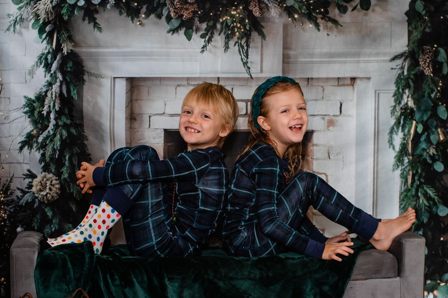 Children in blue plaid pajamas laughing and playing by a festive fireplace setting.