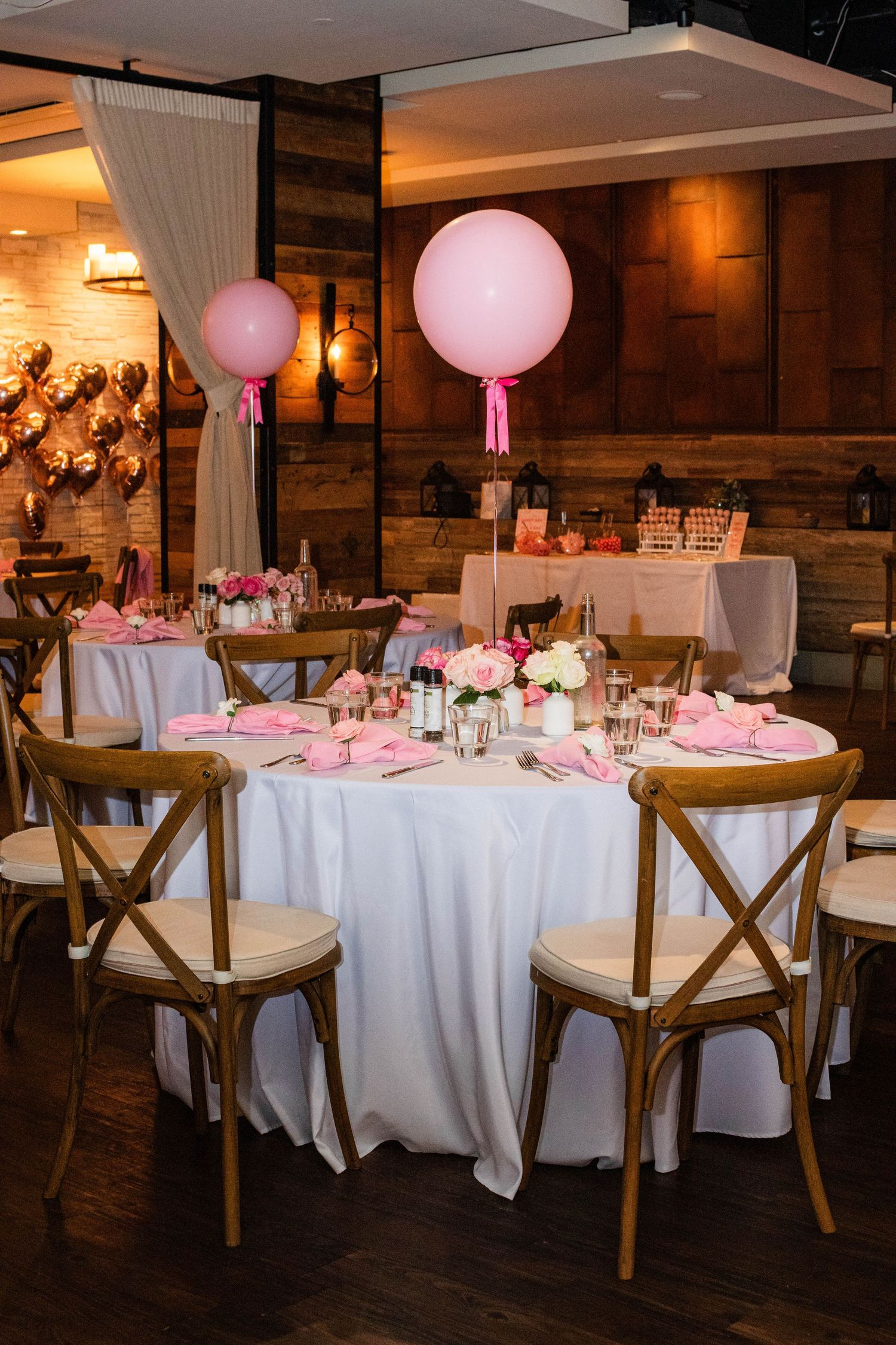 Pink and white decorated wedding reception table with large balloons and wooden cross-back chairs.
