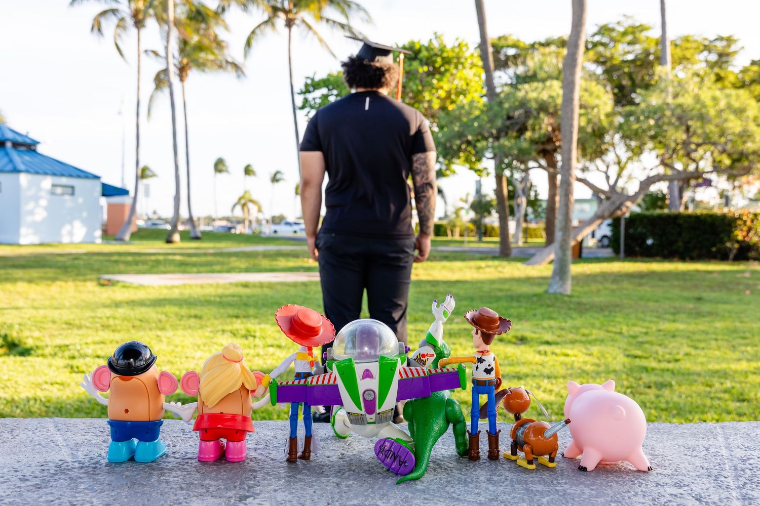 Man in black outfit stands in a park surrounded by Toy Story character figurines including Buzz Lightyear and Woody.