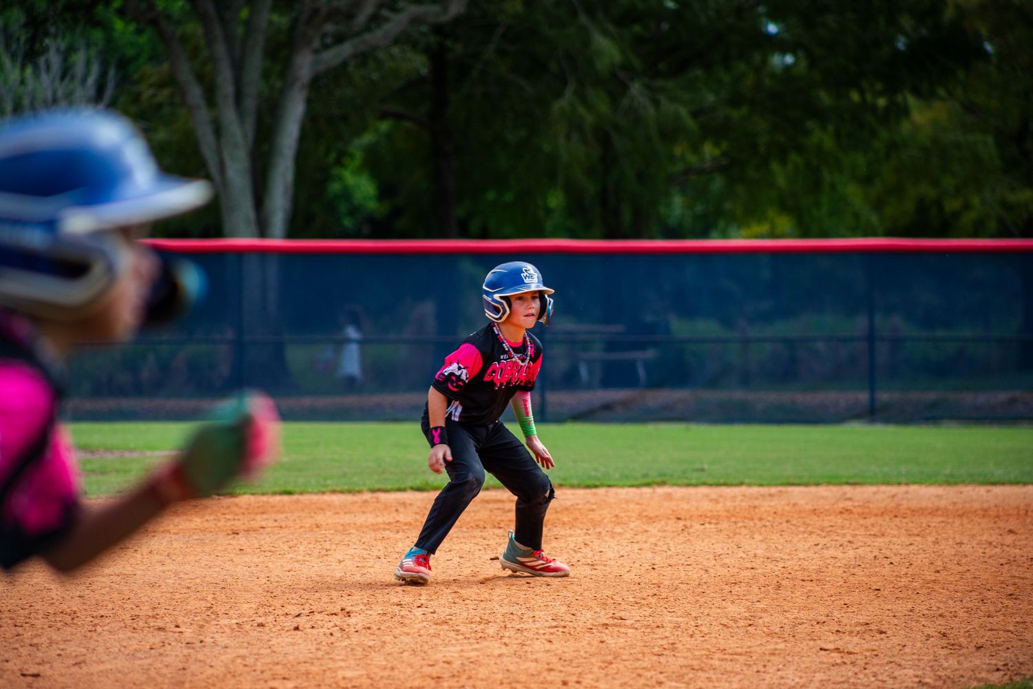 Softball players in action on a dirt infield with trees visible beyond the outfield fence.