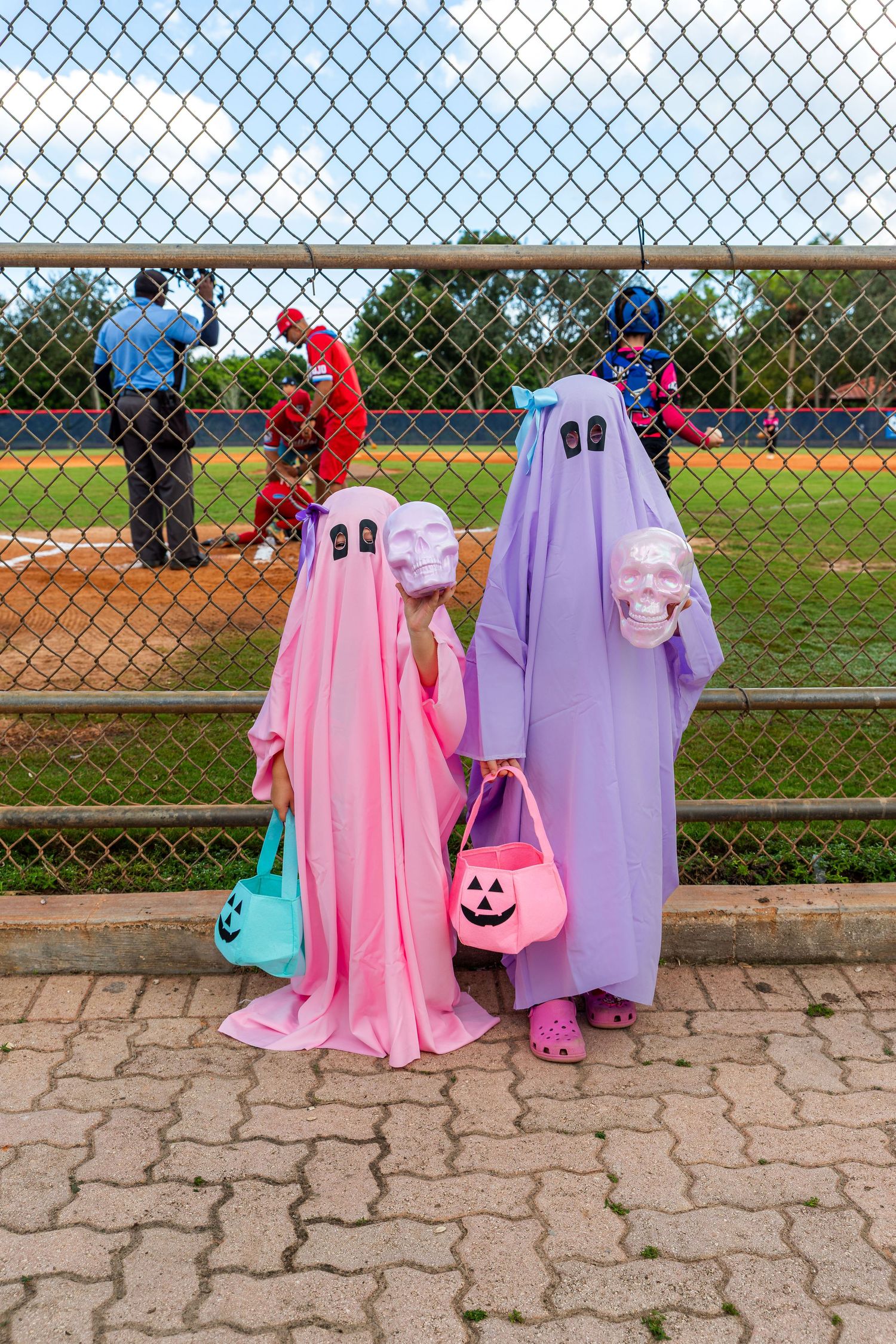 Two people wearing pink and purple ghost costumes stand by a chain link fence at an outdoor event.