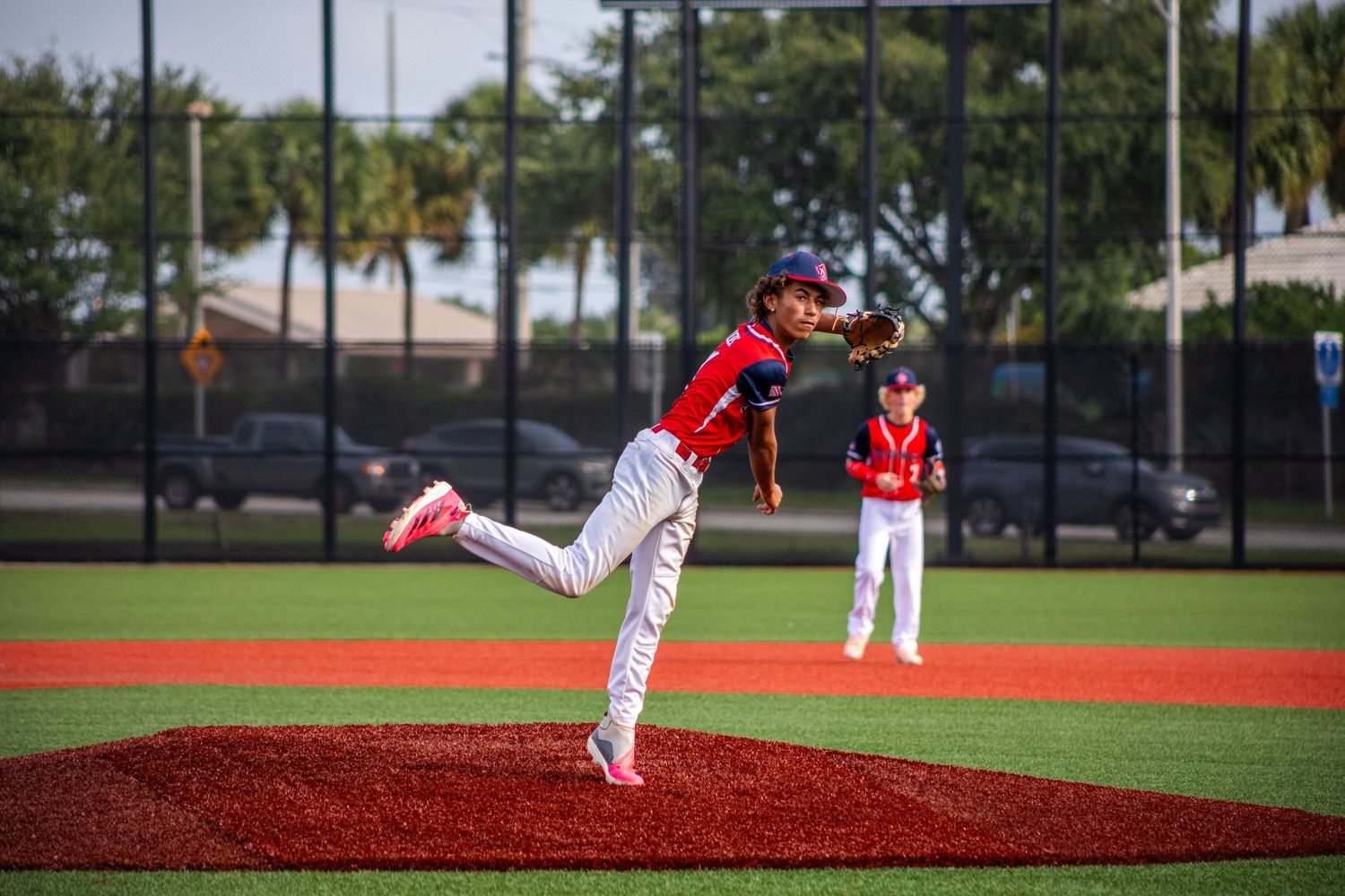 Young baseball pitcher in red uniform delivers a pitch during a youth baseball game on a red turf field.