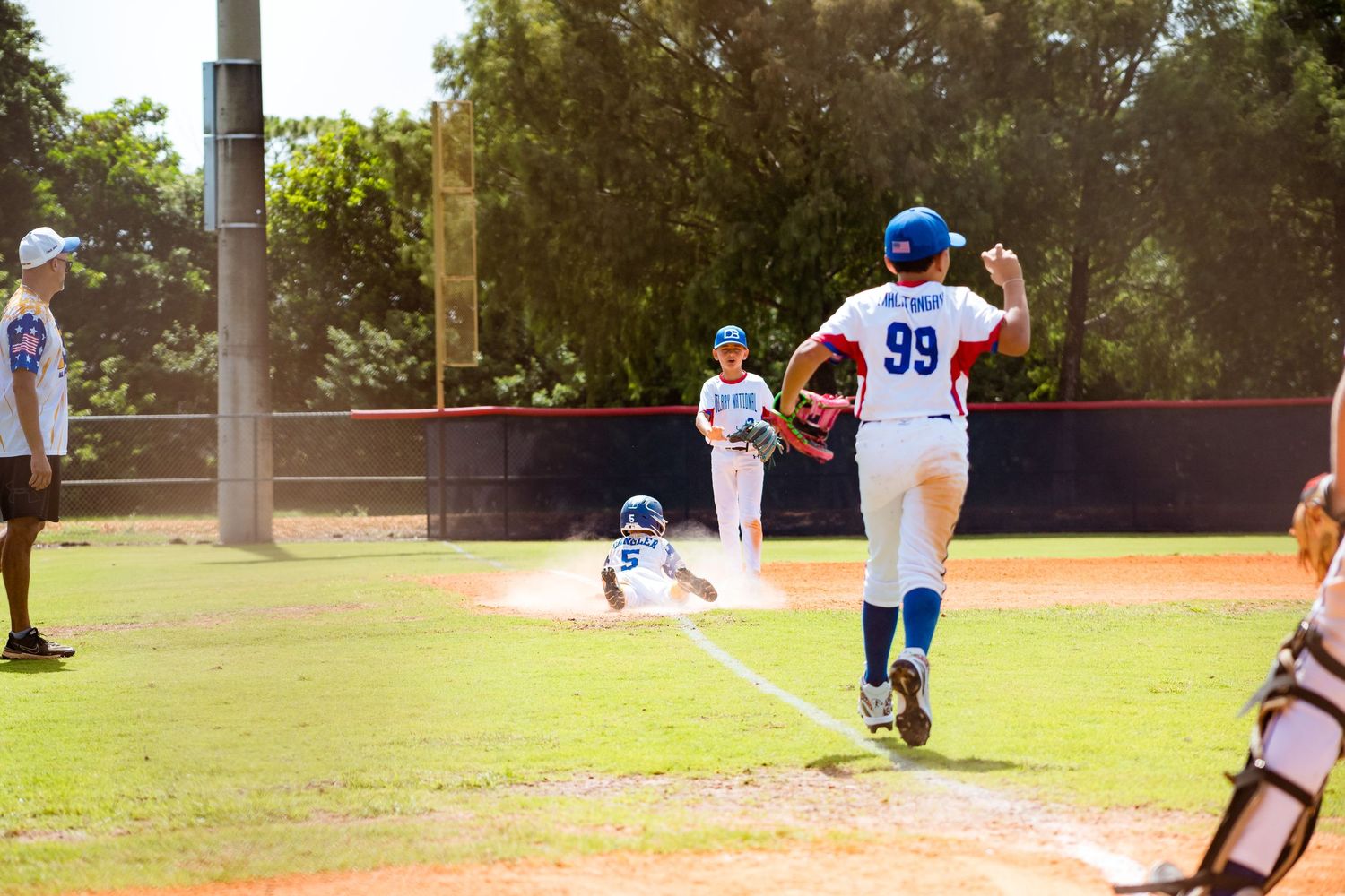 Baseball players spread across a sunny field during a youth baseball game with trees in the background.