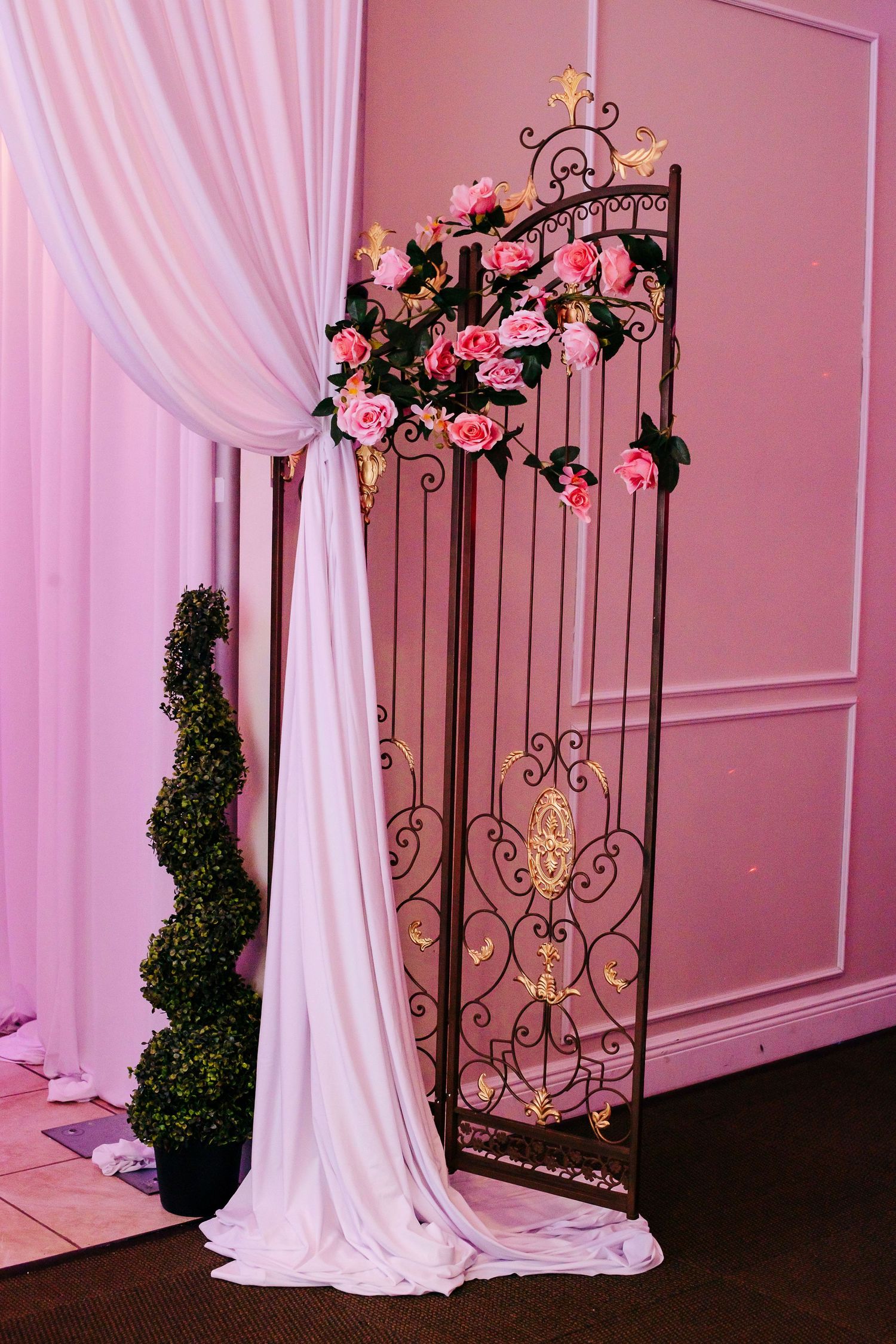 Elegant rose pink decor with romantic iron gates, flowing white drapes and floral arrangements at a wedding venue setup.
