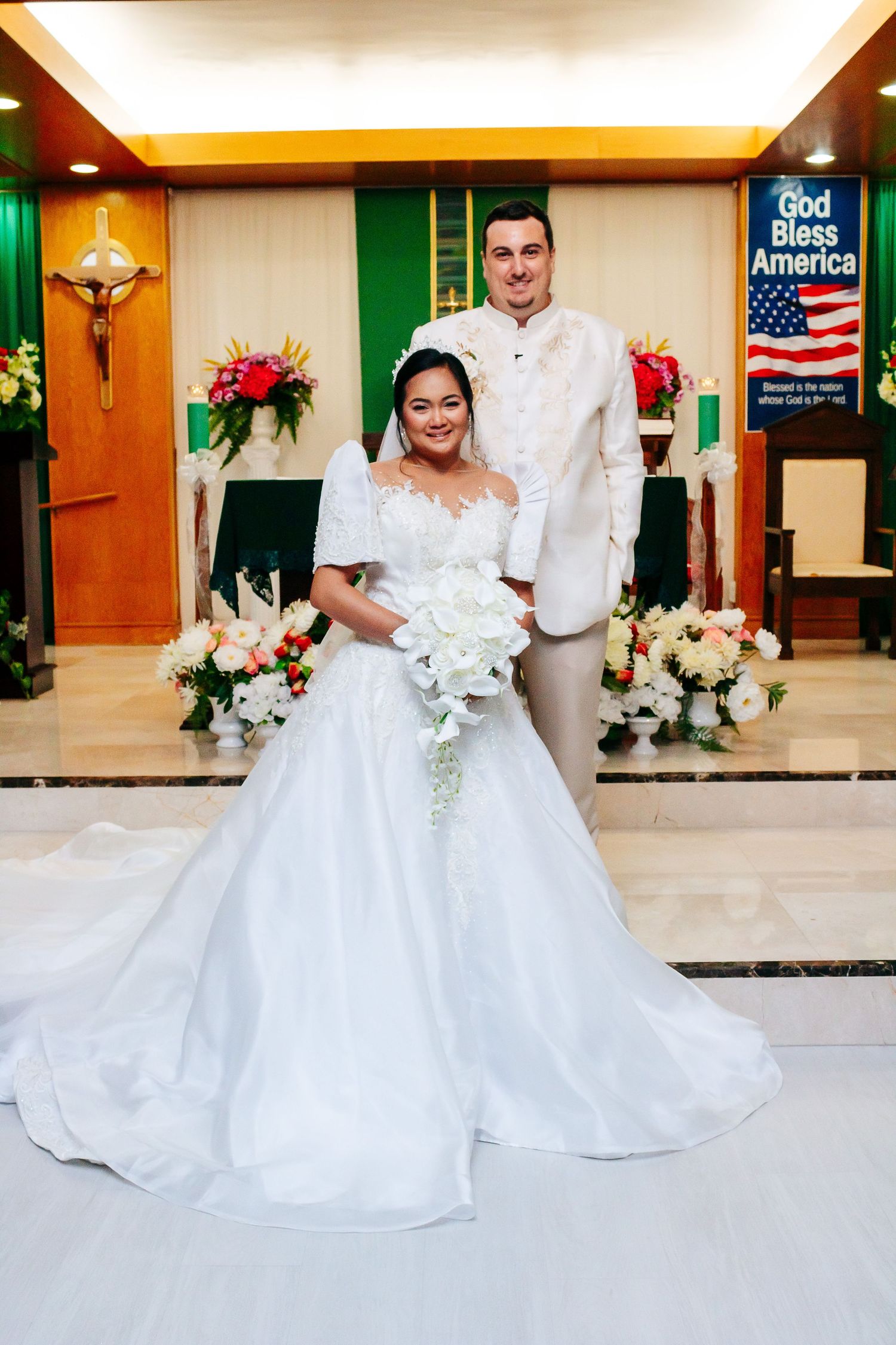 Newlyweds pose for formal portrait in front of church altar with crucifix and flowers.