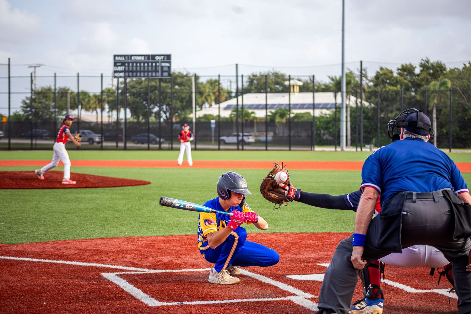 Youth baseball game with batter, catcher, and umpire at home plate, pitcher visible on mound, red turf field.