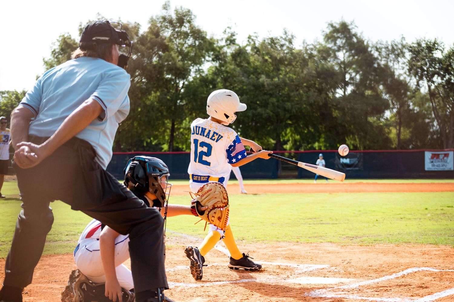 A young baseball player in a white jersey swings a bat at home plate during a youth baseball game.