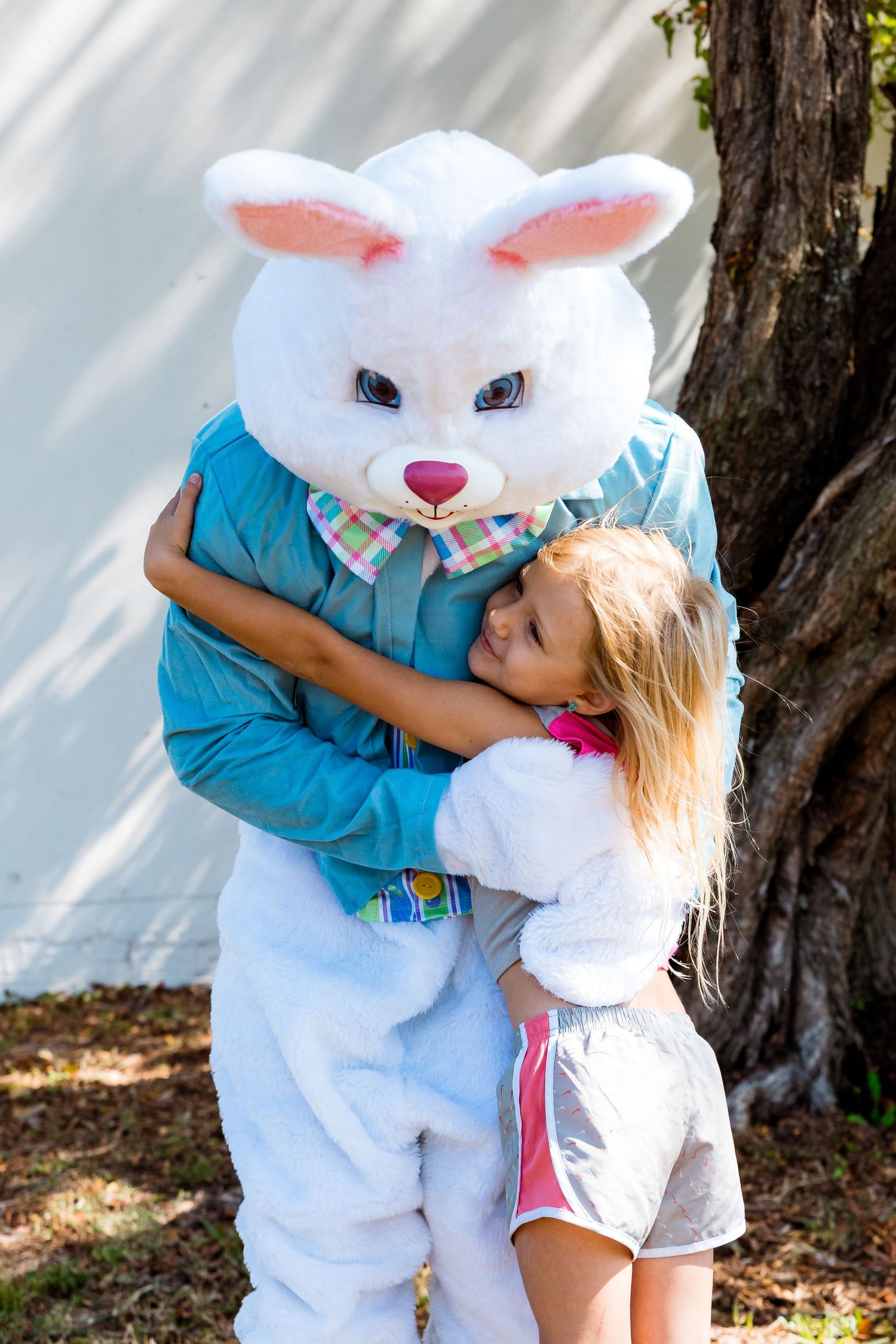 A person hugs a large Easter Bunny mascot wearing a blue shirt and bowtie outdoors near a tree.