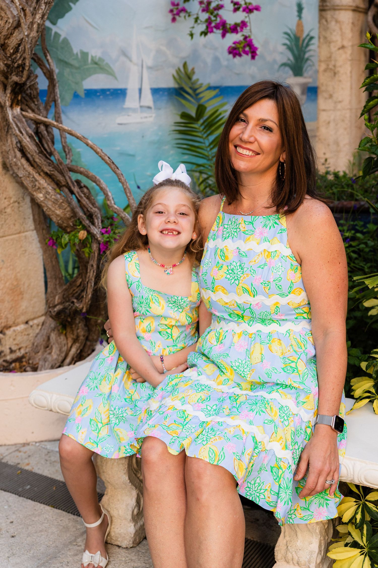 Two people in matching floral print Lilly Pulitzer dresses sitting on stone bench near tropical beach mural.