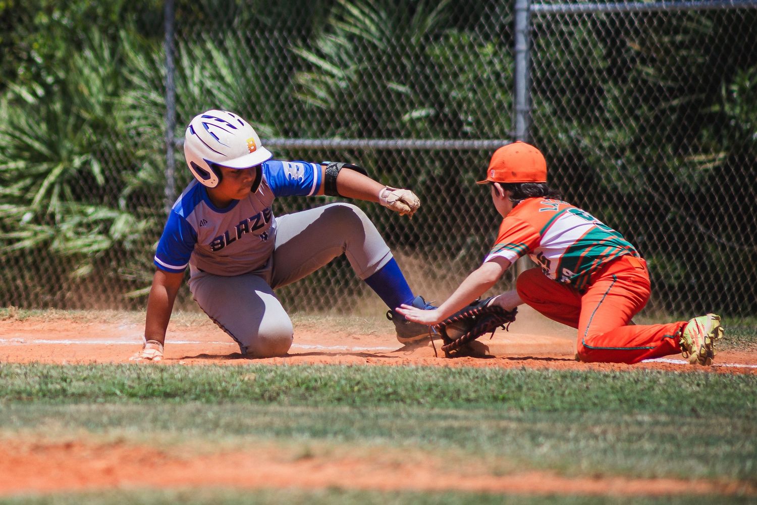 Baseball player in orange uniform attempts to tag runner in blue sliding into base during competitive game on dirt field.