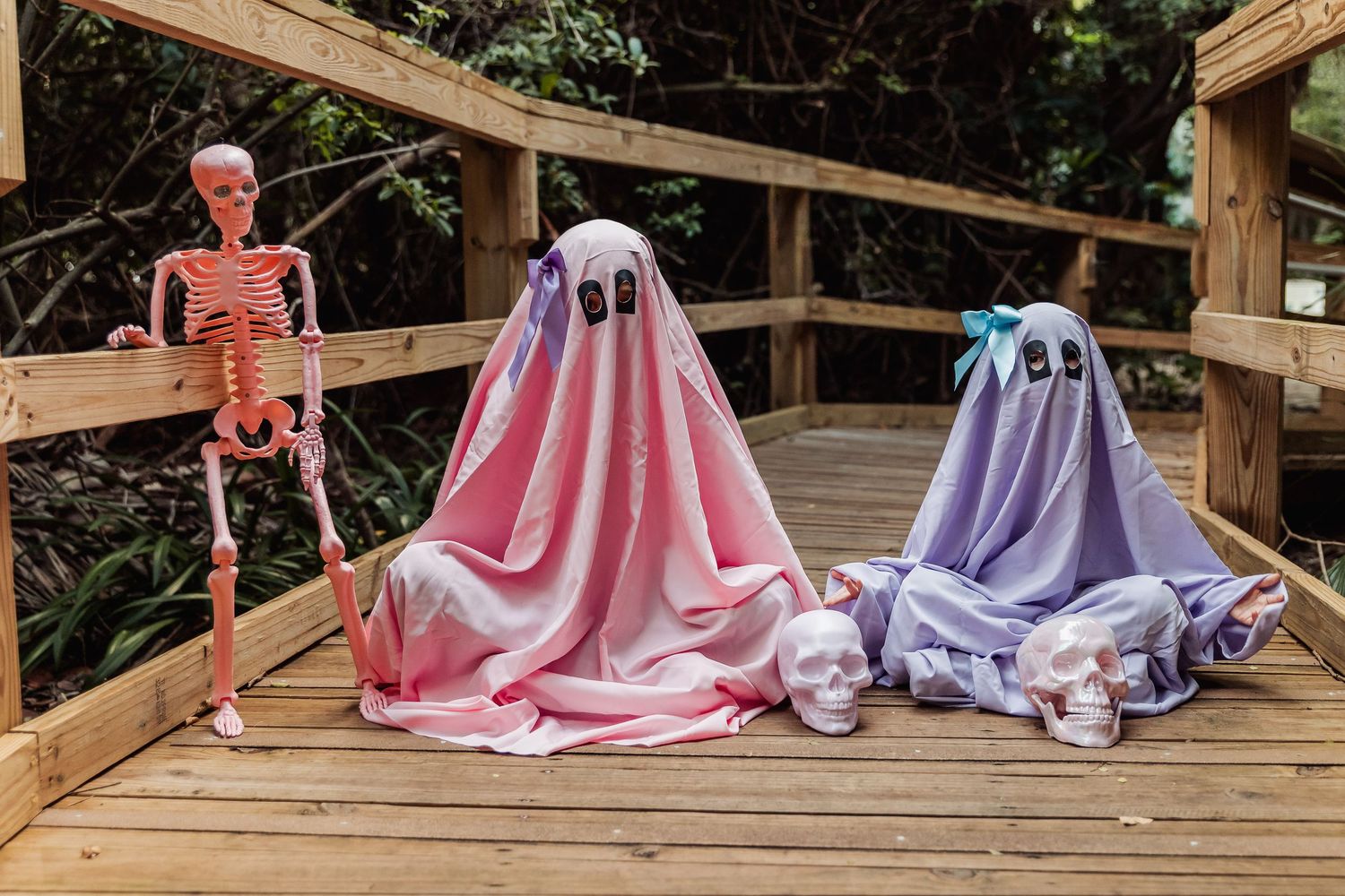 Halloween decorations with a skeleton and two ghost figures draped in pink and purple blankets on a wooden deck.