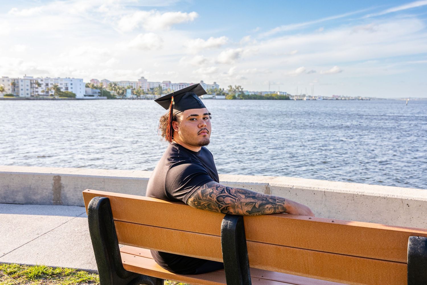 Graduate in cap sitting on bench by waterfront, showing tattoos, with city skyline in background.