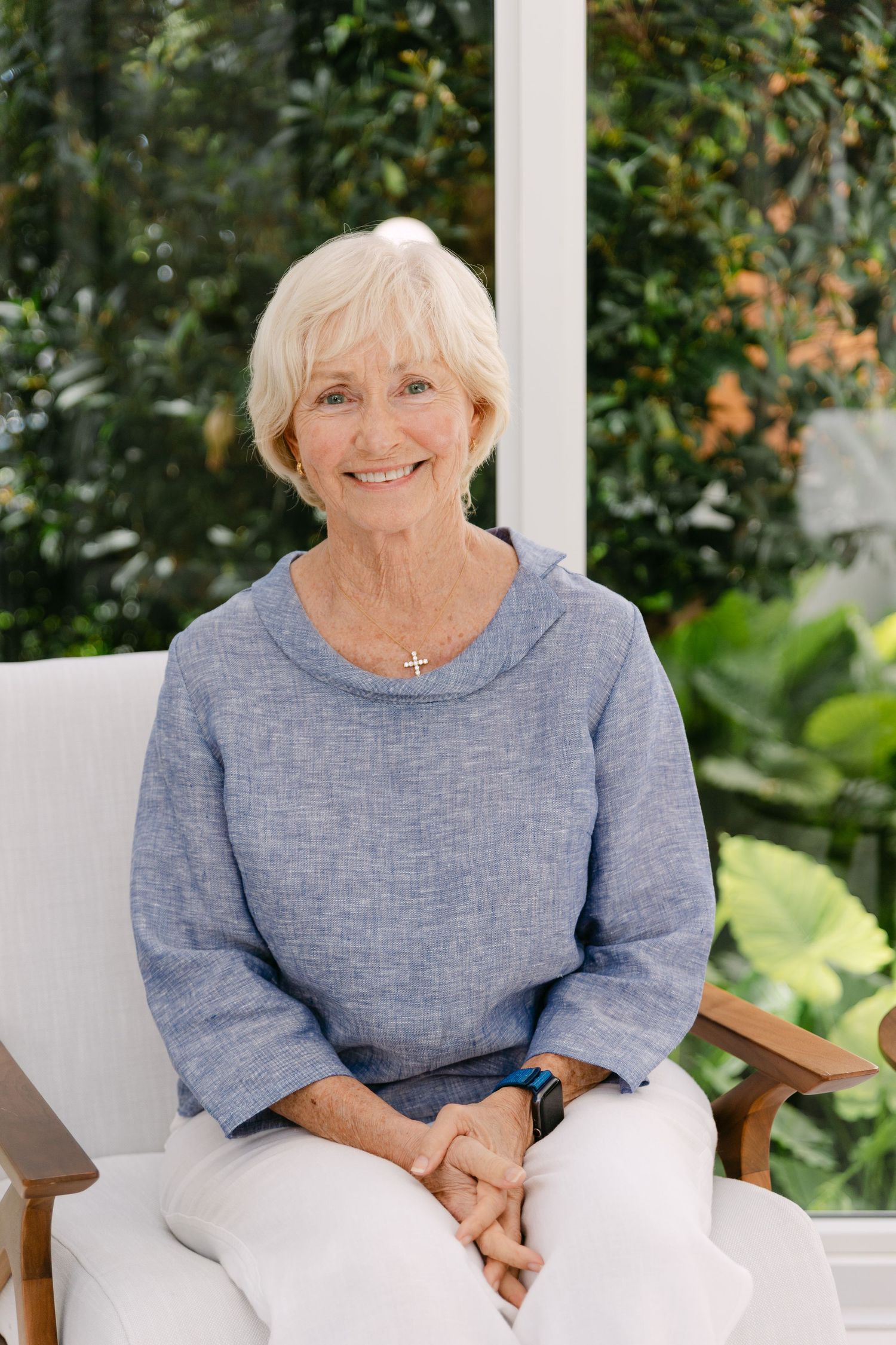 Smiling senior woman with white hair sitting in a chair, wearing a blue linen top and white pants outdoors.