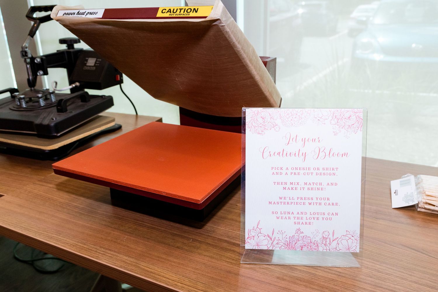 A heat press machine and pink invitation card on a wooden desk.