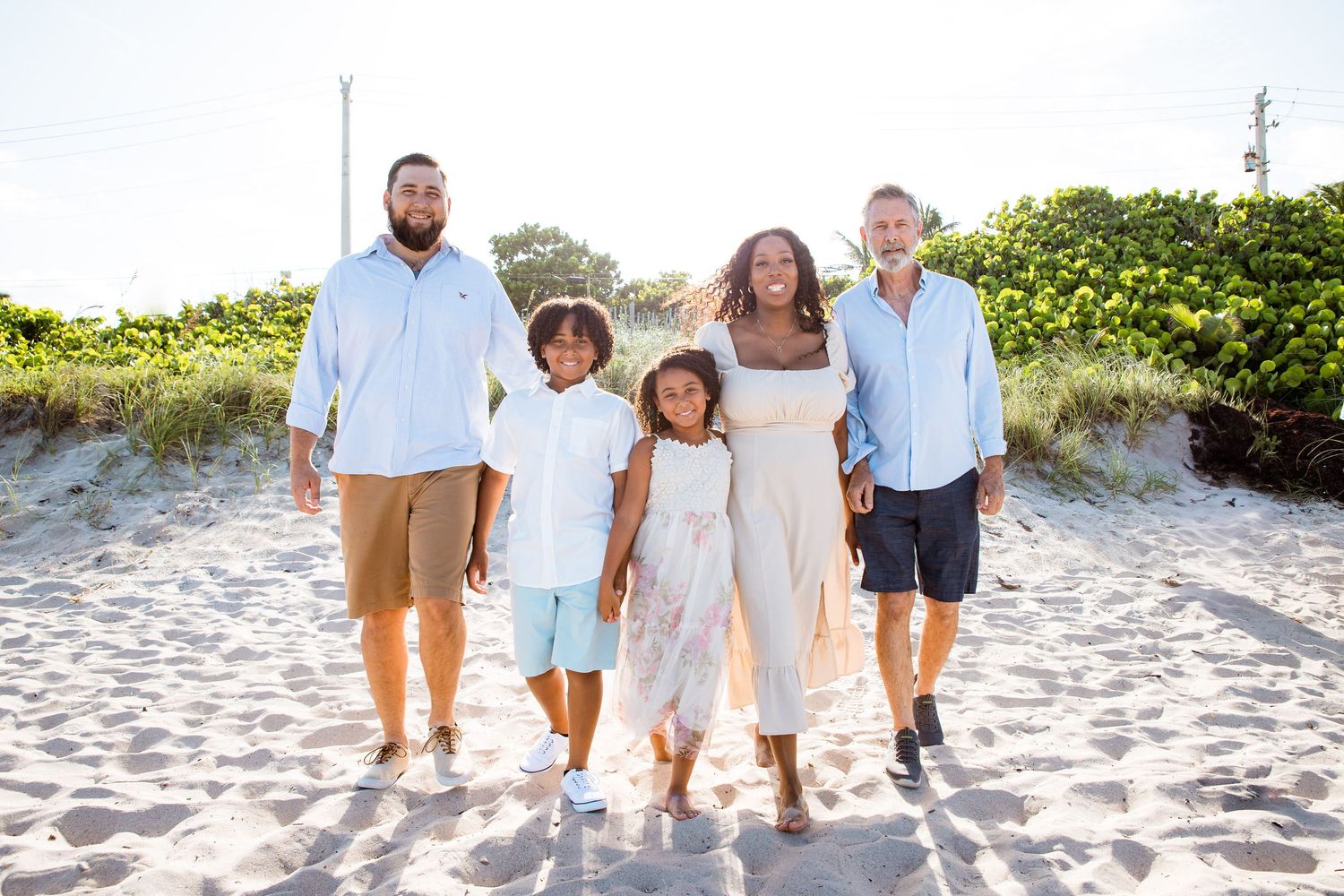 A group of five people walking together on a sunny beach with sand dunes and greenery in the background.