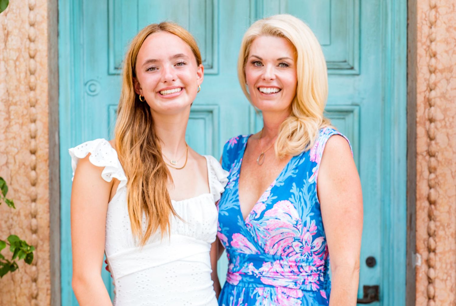 Two people in summer attire smile brightly while standing in front of a turquoise blue door.